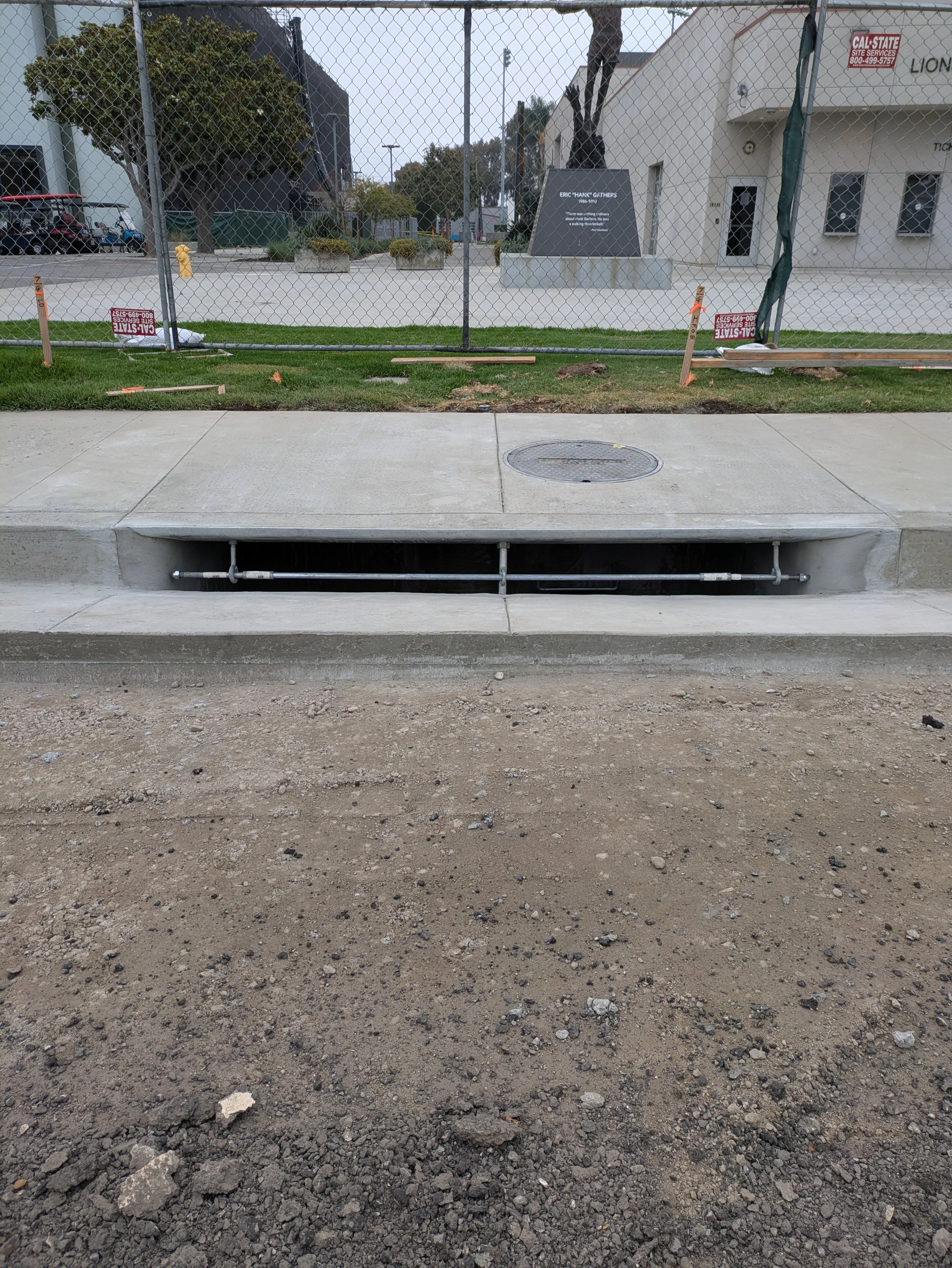 View of a sidewalk with a nearby underground utility access panel and exposed pipes, asphalt and dirt ground, behind a temporary chain-link fence, with construction warning signs. In the background, there are buildings, a tree, and signs related to s