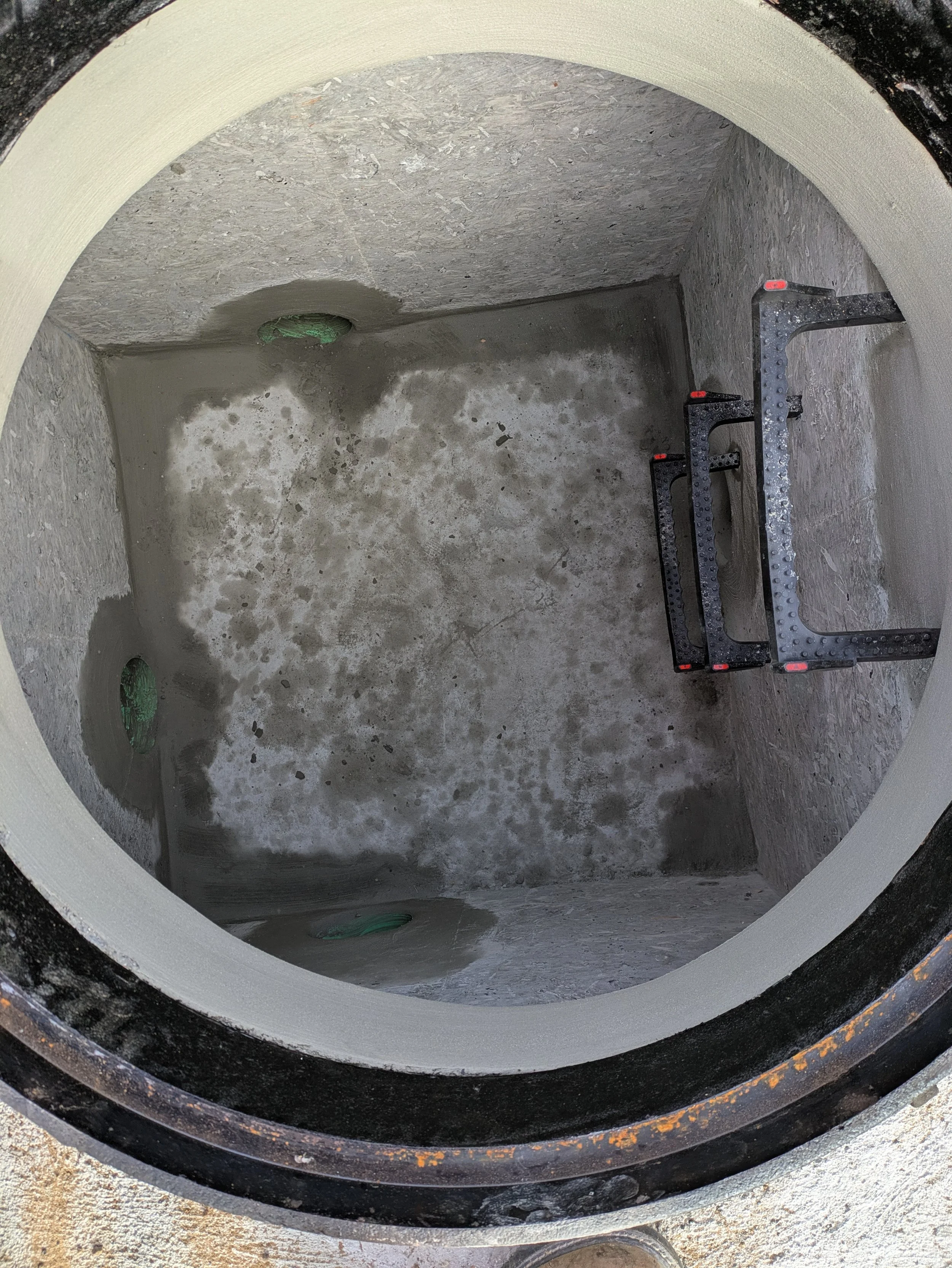 The interior of a cylindrical well with concrete lining, showing construction materials and tools including black metal brackets with red caps and green plastic components.