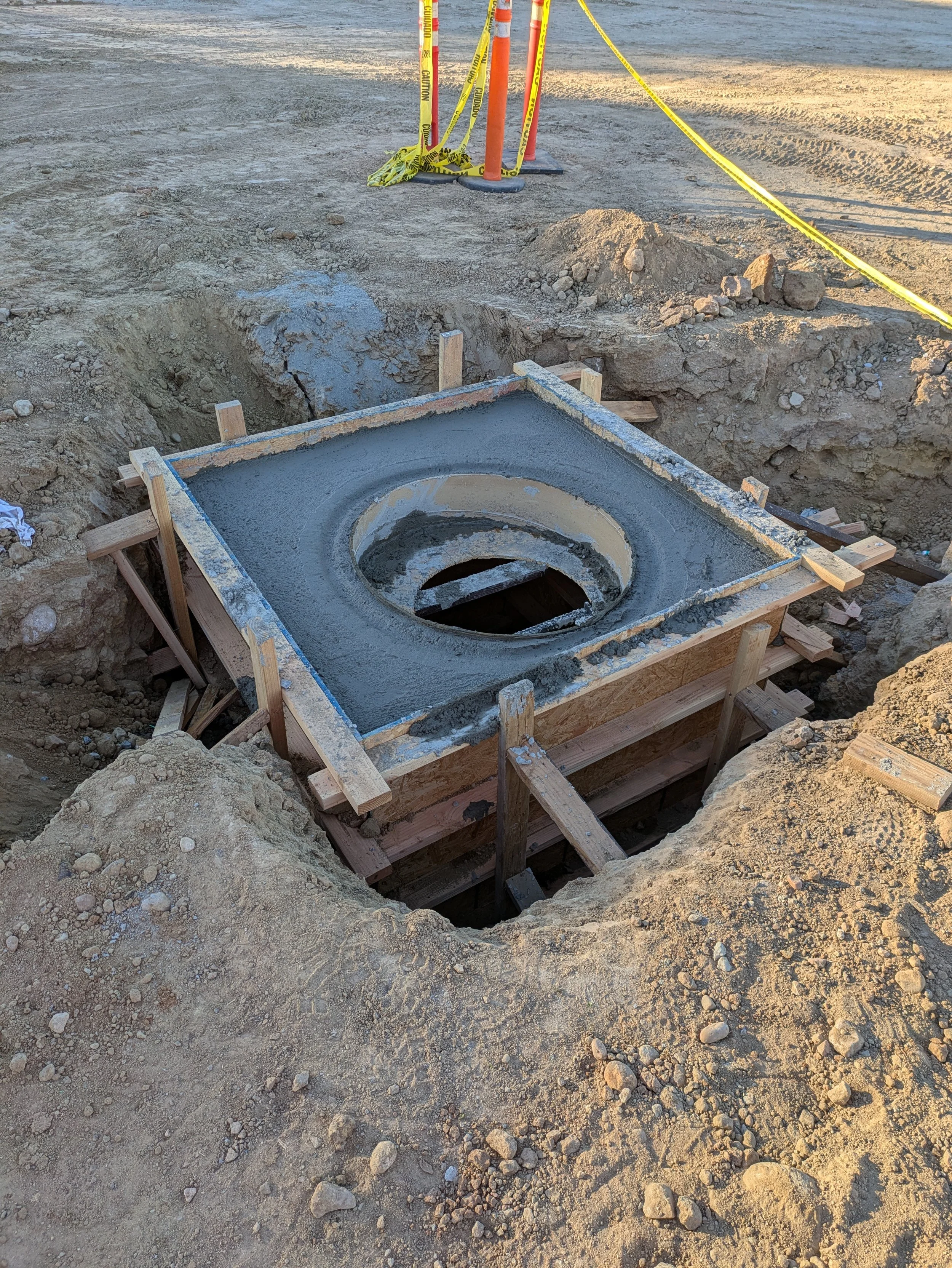 Construction site with a freshly poured concrete circular base in a dug-out hole, surrounded by dirt and loose rocks. Yellow caution tape and orange cones mark the area.