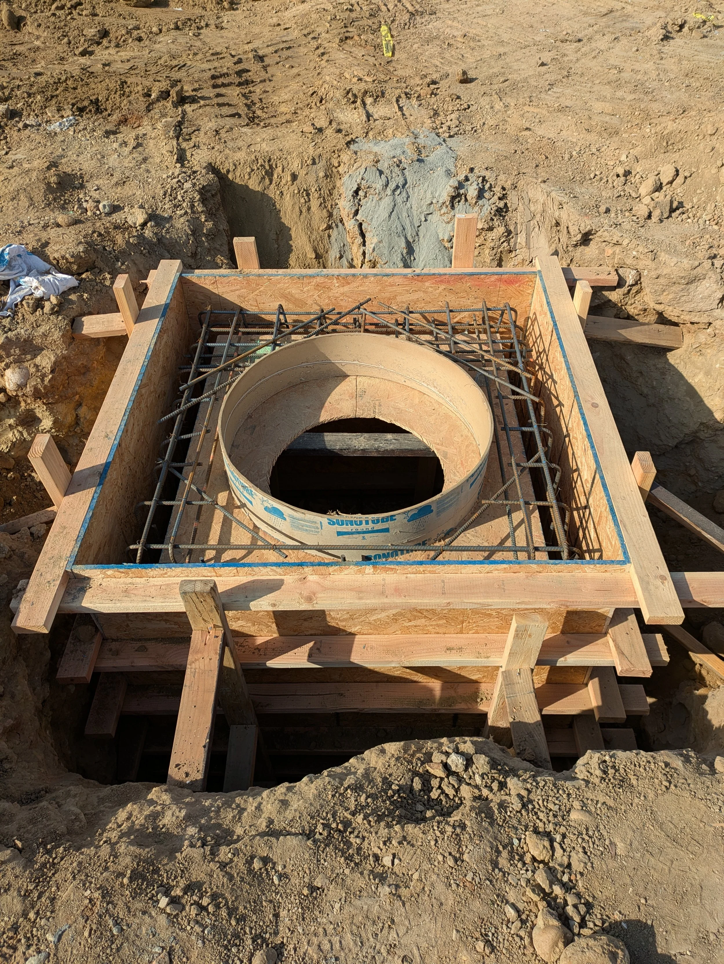 Construction site with a concrete pipe in a wooden formwork, surrounded by dirt and rocks, for underground utility or drainage installation.