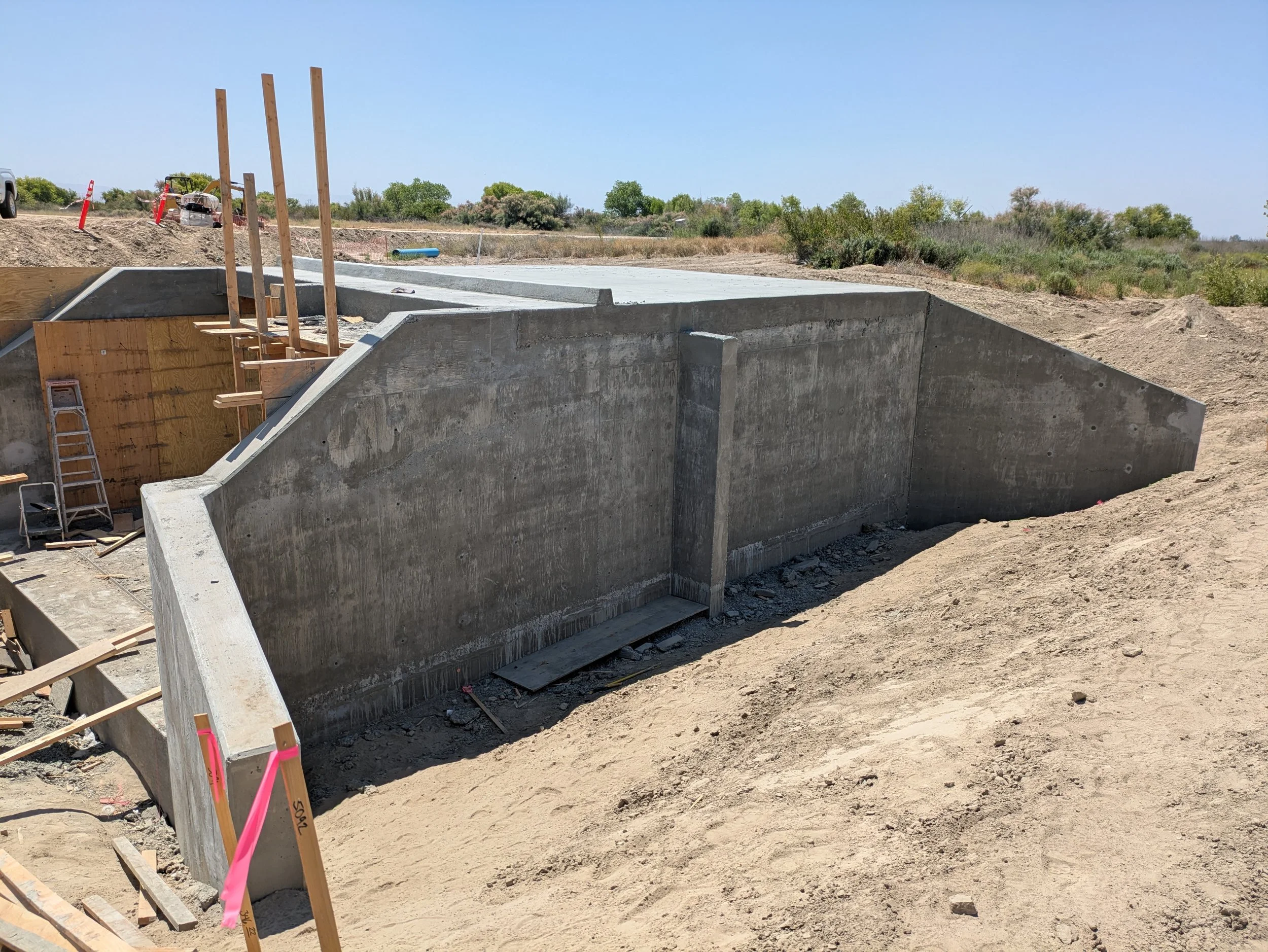 Construction site with concrete foundation walls and wooden formwork in progress, surrounded by dirt and construction materials under a clear blue sky.