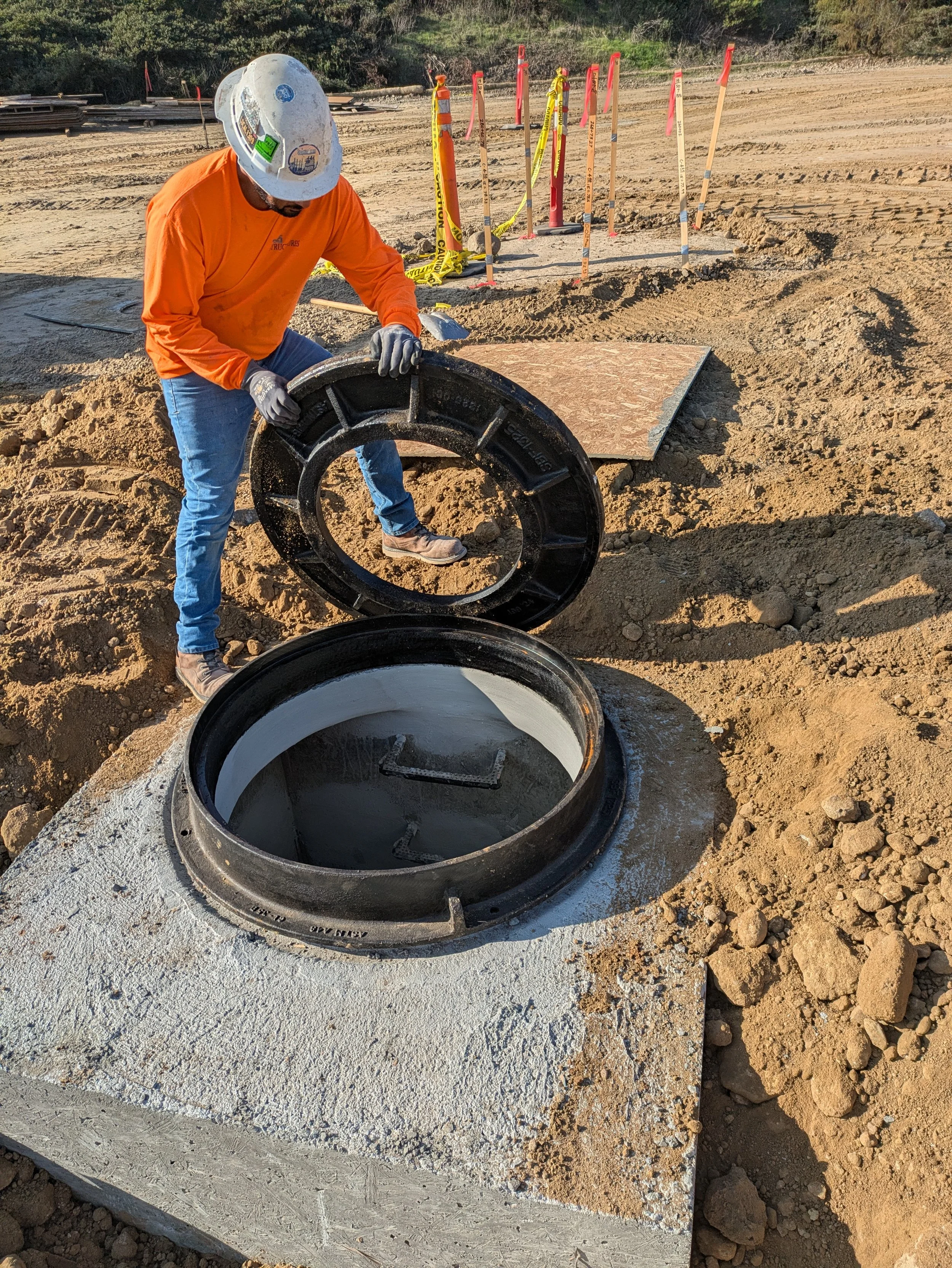 A construction worker in a hard hat and orange shirt installing a large concrete utility ring into the ground at a construction site with safety marked poles in the background.