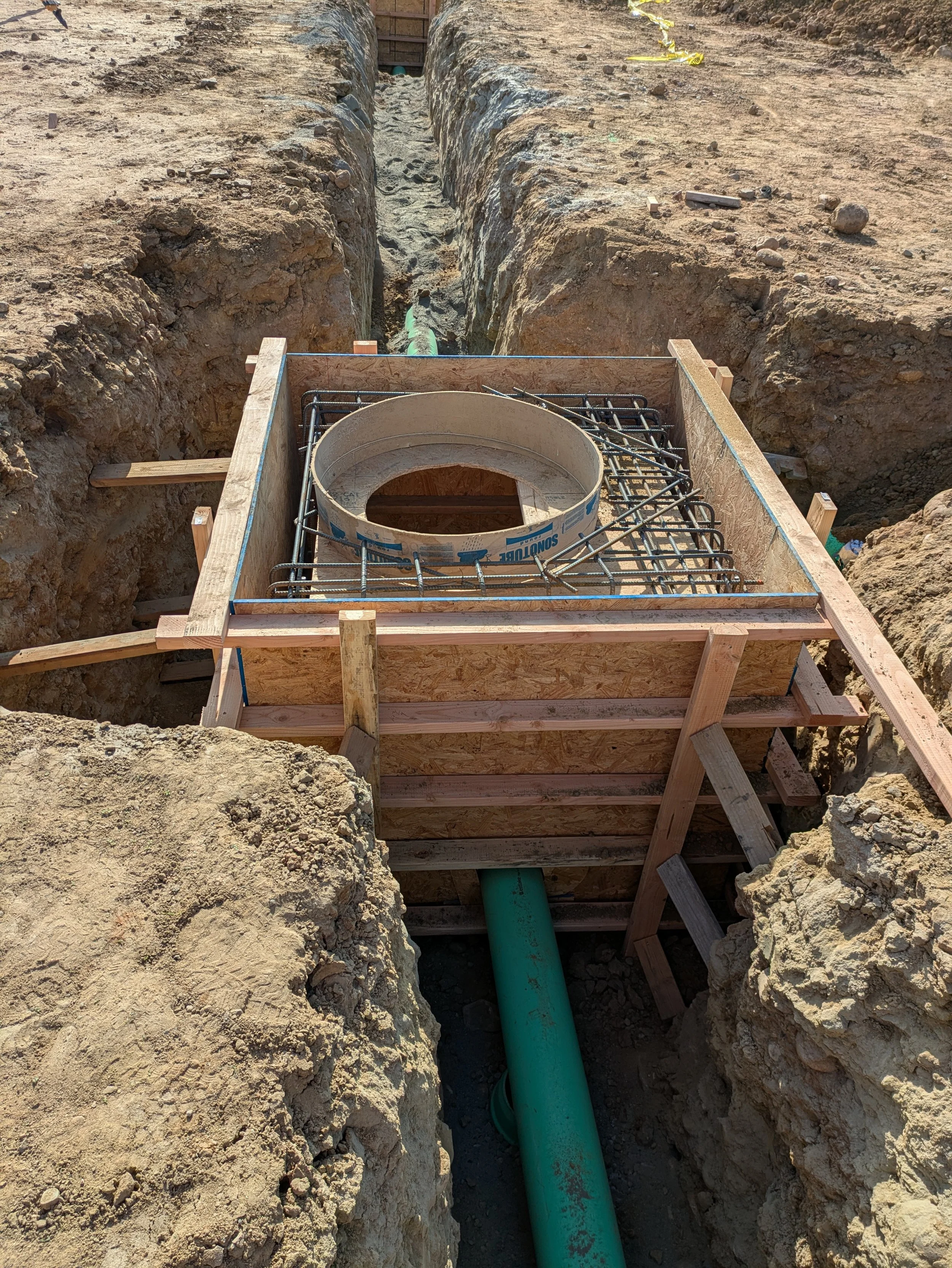 Construction site with dug trench and green underground pipe, with wooden formwork and rebar reinforcement around the pipe.