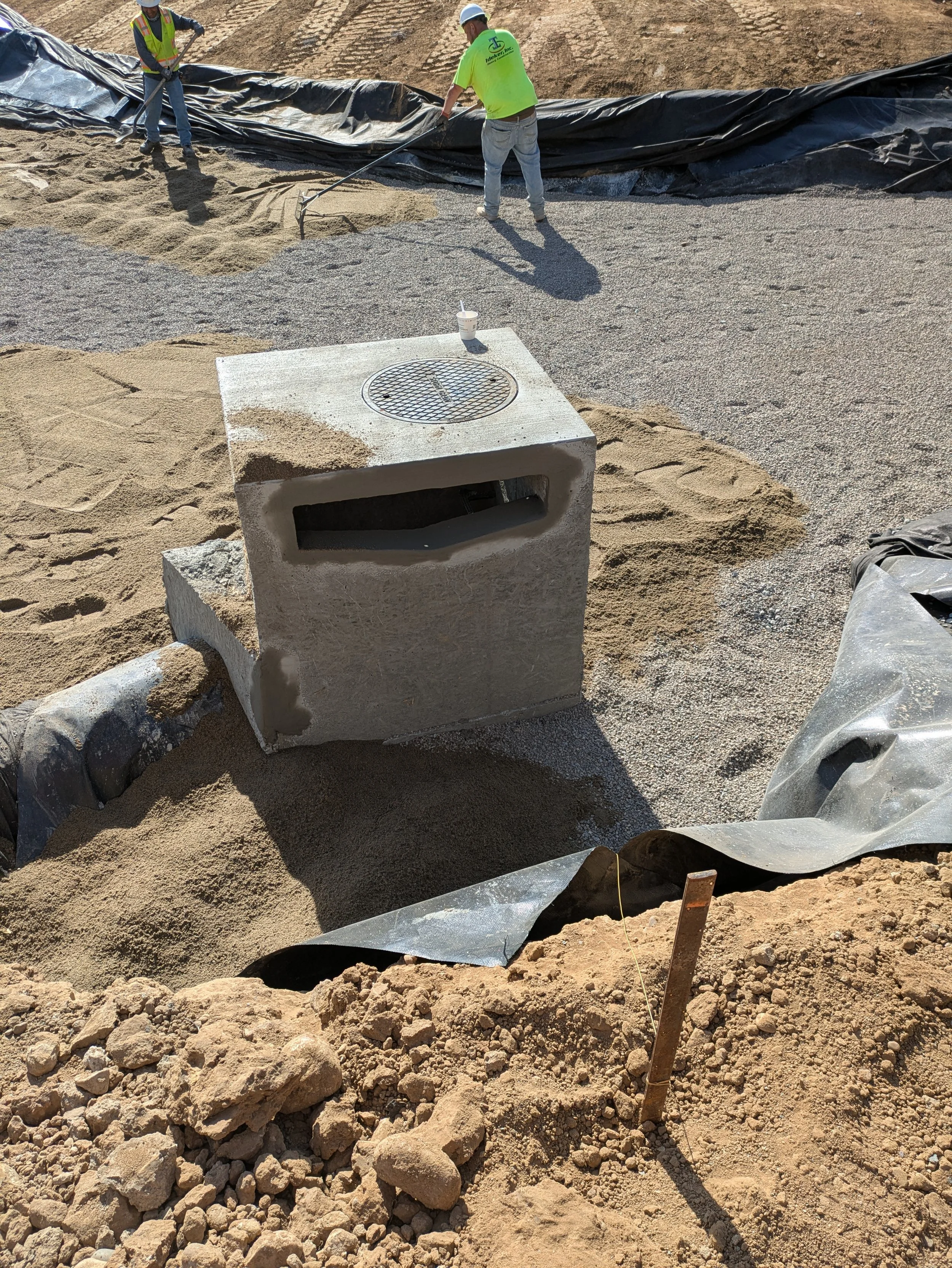 Workers in safety gear involved in construction work on a reddish-brown dirt site, laying down gravel or similar material with black plastic sheeting in the background.