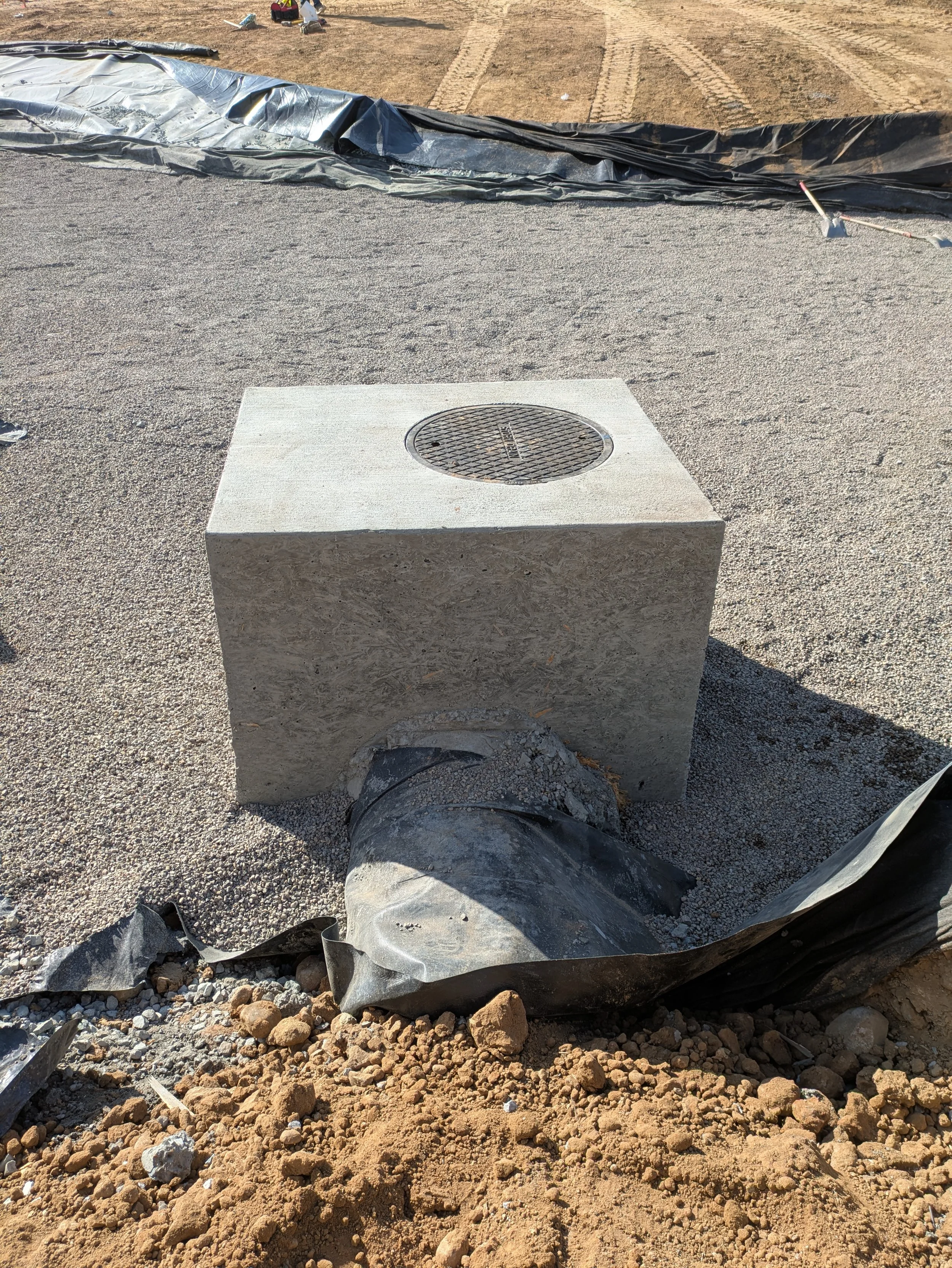 The image shows a construction site with a partially buried manhole cover, surrounded by gravel and dirt, and a large tarp or plastic sheet partially covering the area.