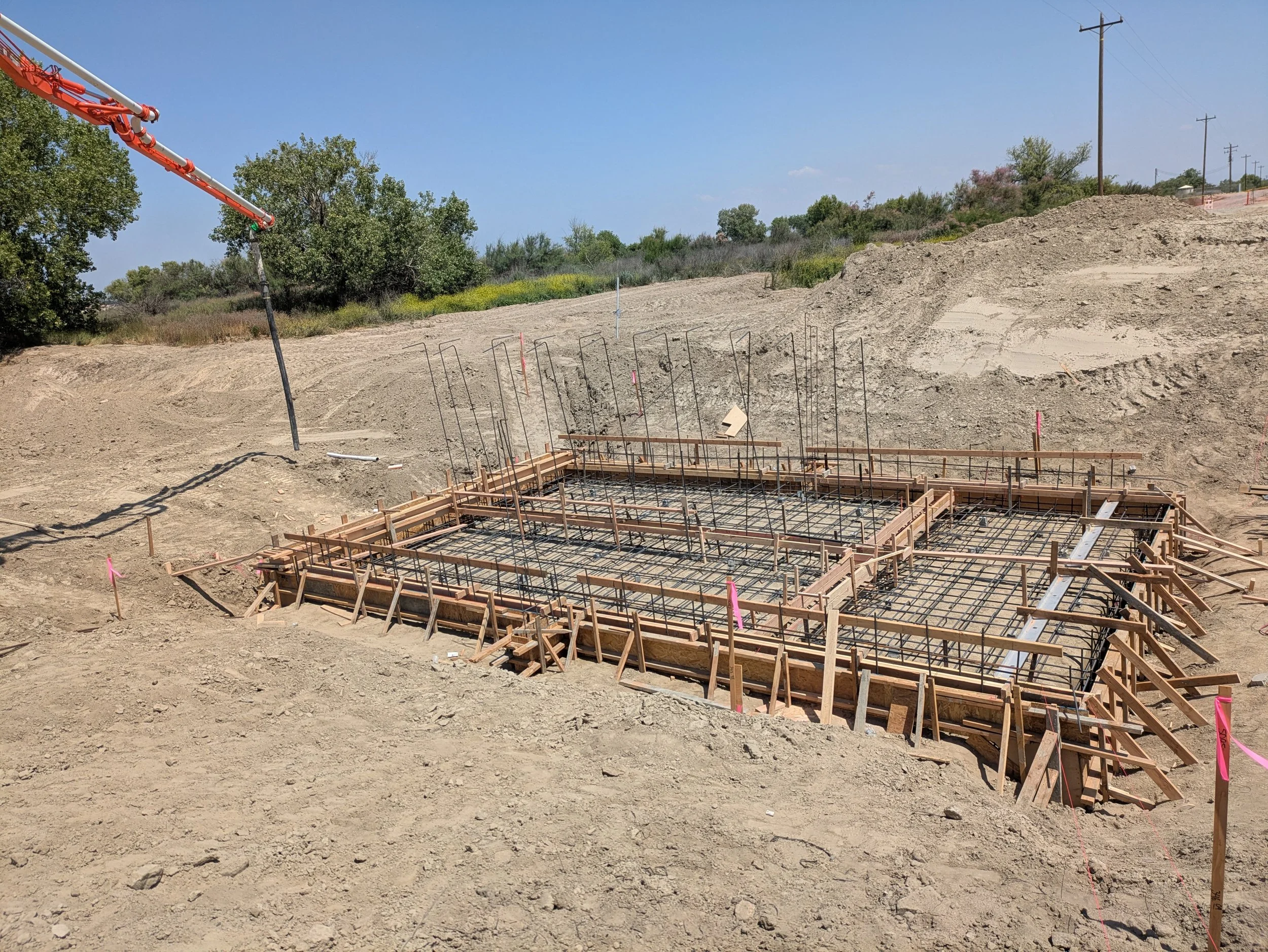 Construction site with concrete foundation framing, dirt mounds, trees, utility poles, and a clear blue sky.