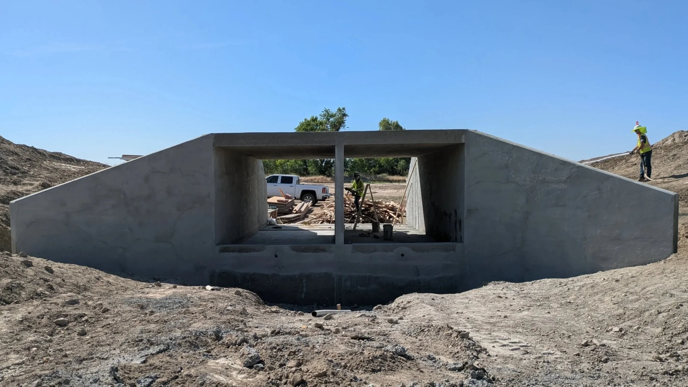 Construction site with a concrete structure and workers in safety gear, surrounded by dirt mounds, a pickup truck, and trees in the background under a blue sky.