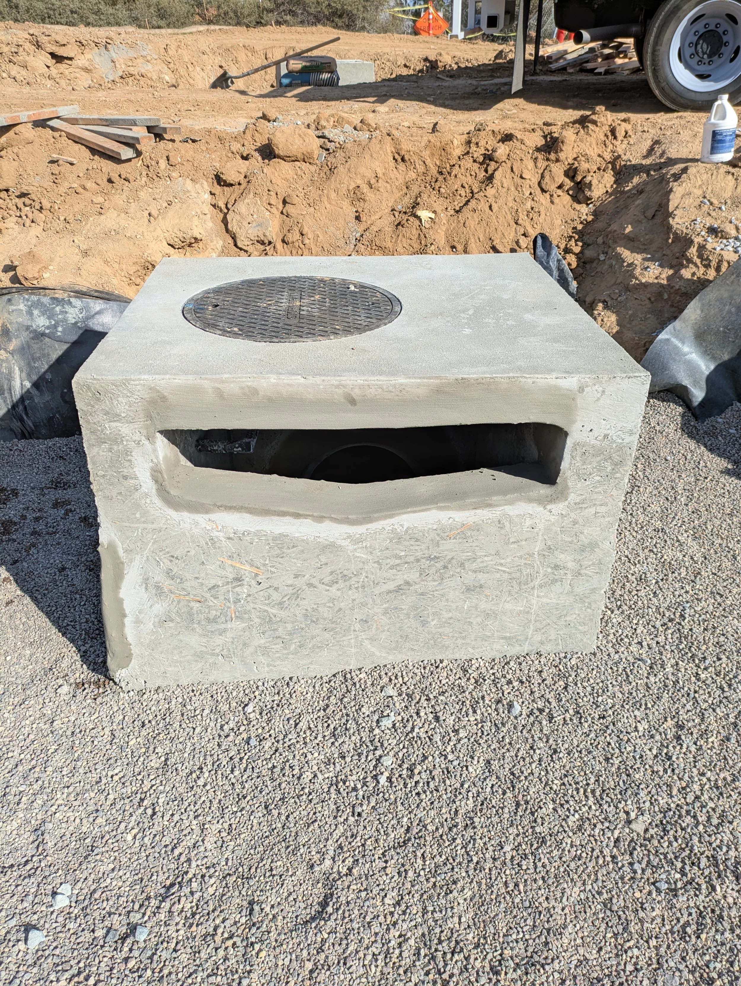 A construction site with a concrete block that has a round metal cover on top, surrounded by dirt, gravel, and construction tools.