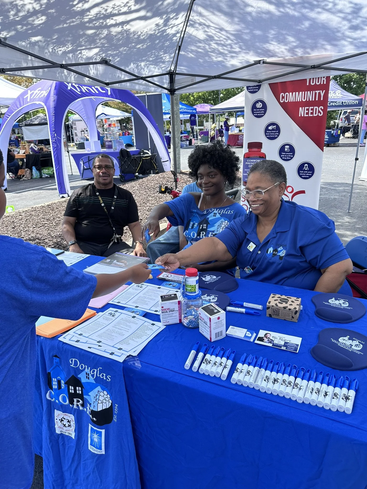Community outreach booth with informational materials, pens, and medication bottles, under a white canopy, with people sitting and standing nearby at an outdoor event.