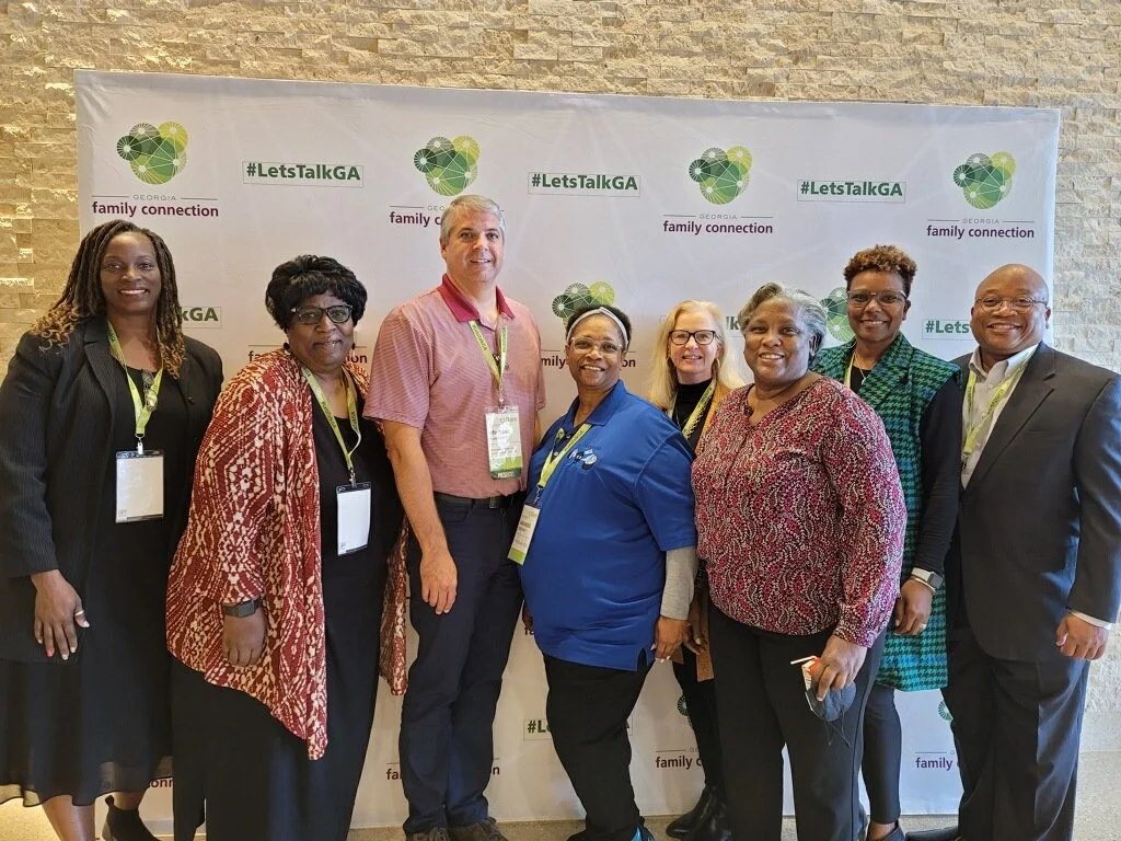 Group of eight diverse people standing in front of a backdrop with logos for Georgia Family Connection and the hashtag #LetsTalkGA, smiling at the camera.