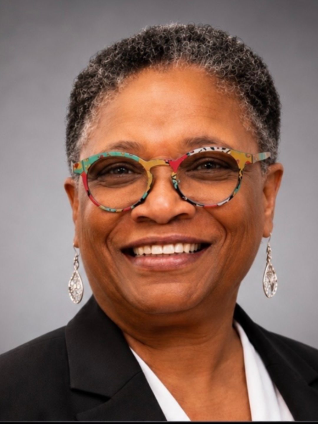 Close-up of Amanda Bryant wearing colorful glasses, earrings, black blazer, and a white shirt against a plain gray background.