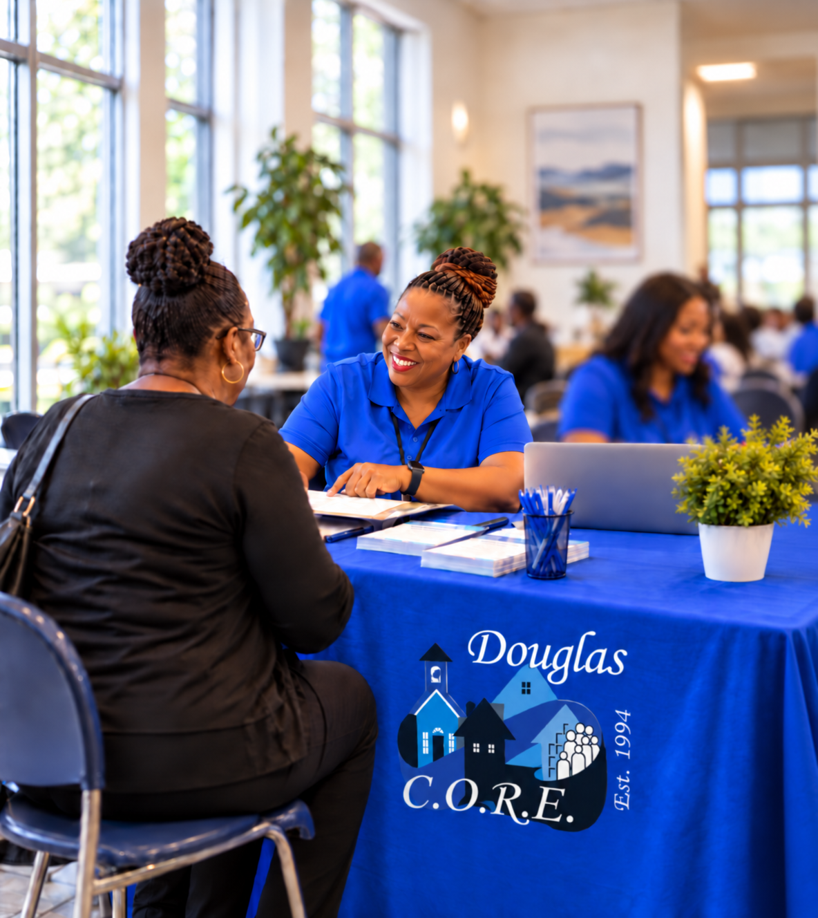 Two women sitting at a table engaging in conversation inside a bright, airy room with large windows. One woman is wearing a black top and glasses, and the other is wearing a blue shirt and smiling. The table has a blue tablecloth with the logo 'Douglas C.O.R.E. Est. 1994,' a potted plant, notebooks, and a laptop.
