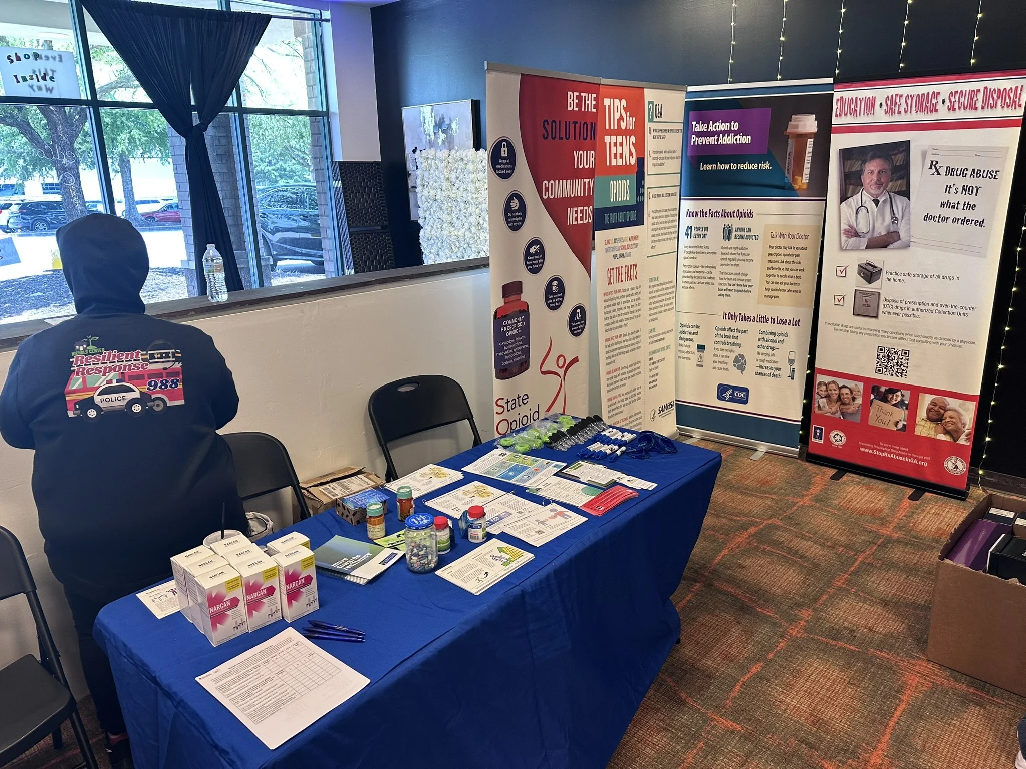 An informational booth with various brochures and medication samples related to drug addiction and opioid awareness. There are informational posters, a table with brochures, and a person wearing a jacket with a police emergency contact number at the back.