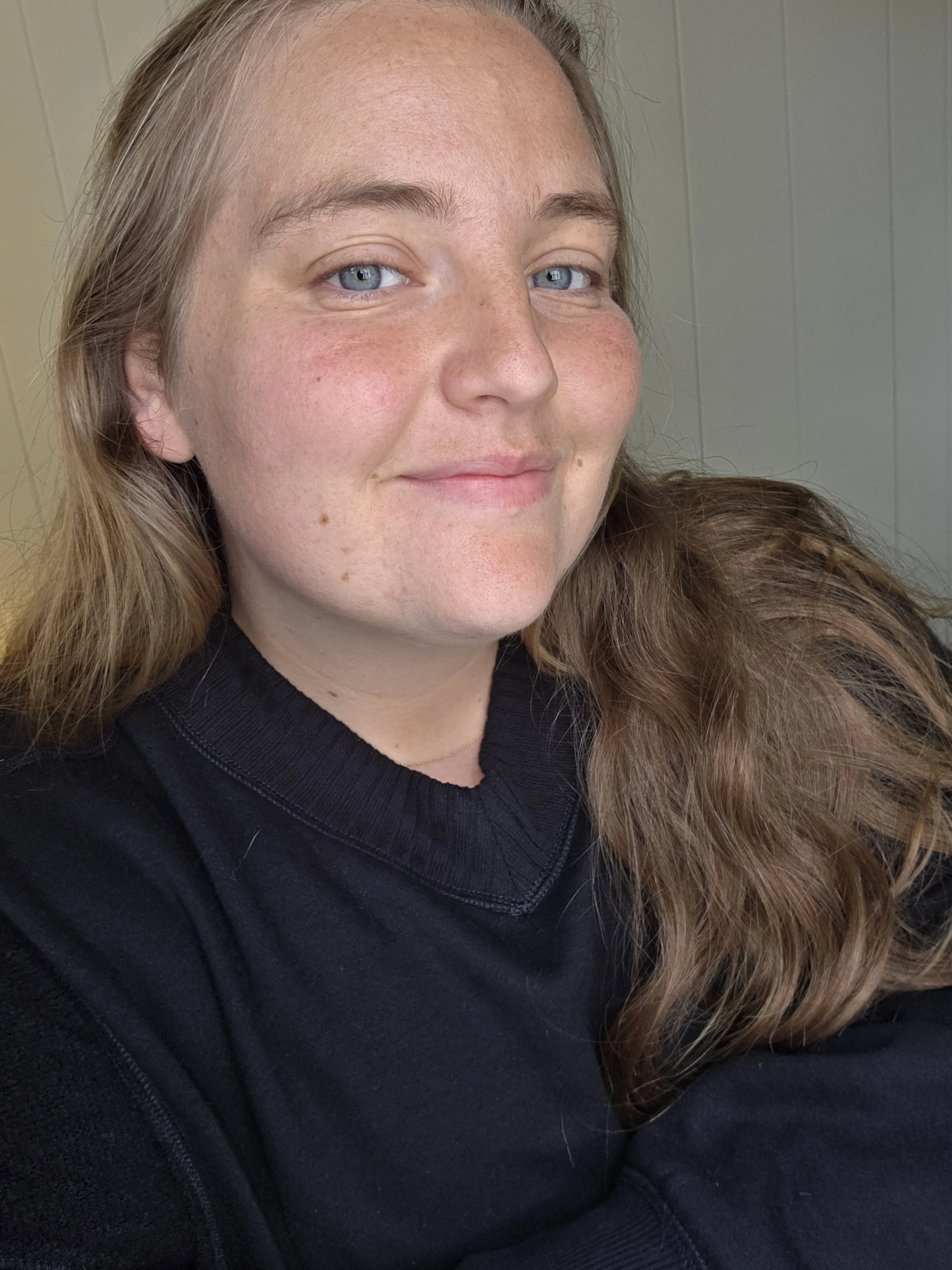 A close-up of a woman with long, wavy light brown hair and blue eyes, smiling slightly. She is wearing a black shirt and the background shows a beige wall.