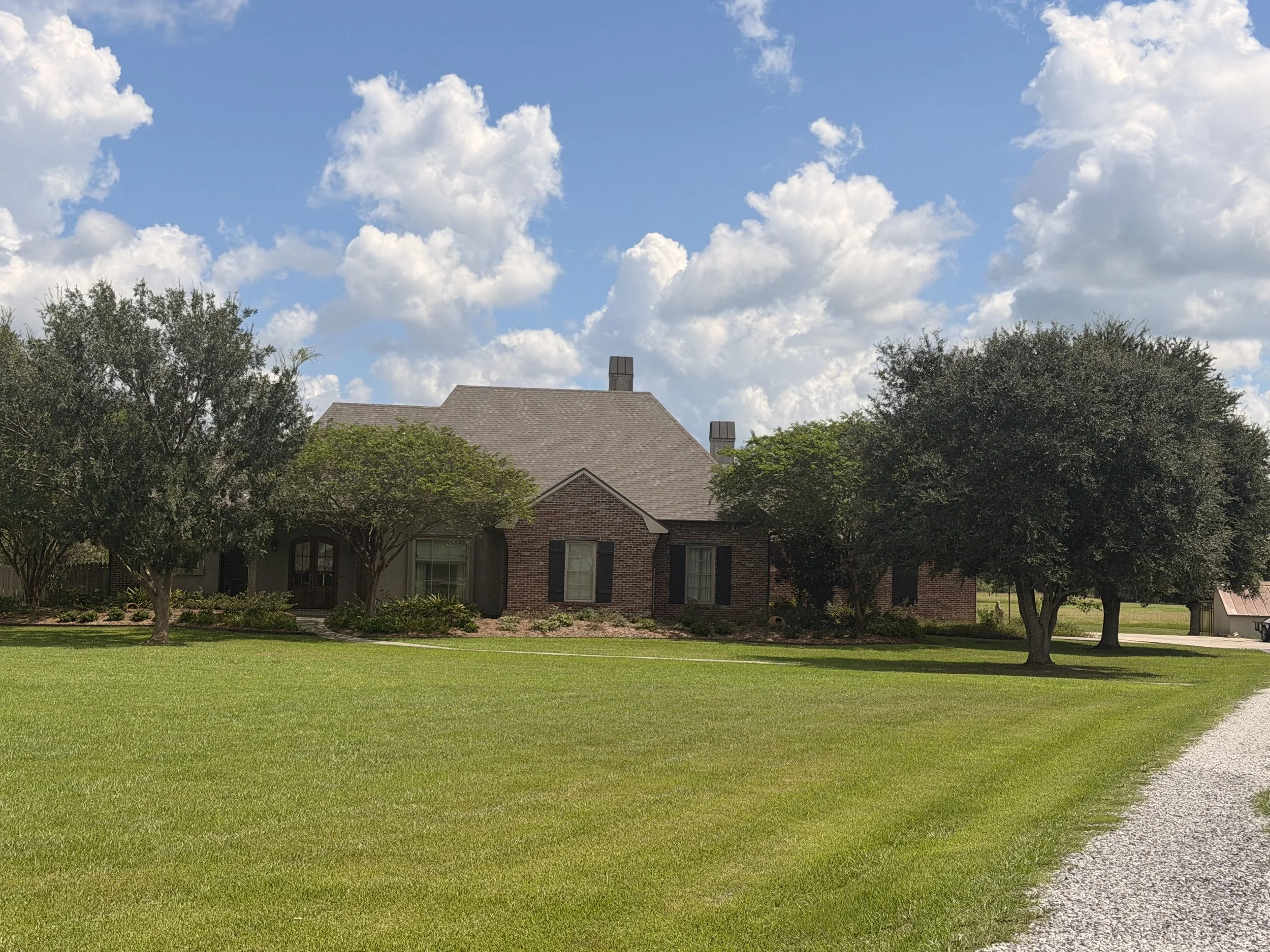 A house with a brick exterior, surrounded by trees, under a blue sky with white clouds and a grassy lawn in the foreground.
