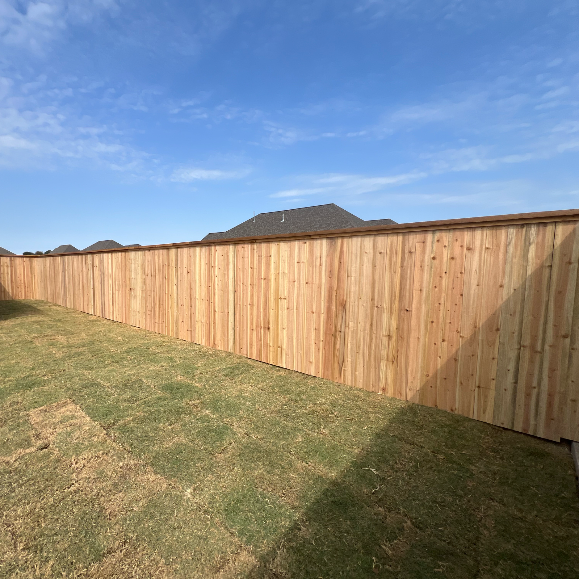 A wooden privacy fence with a brownish color, situated on a grassy lawn, with the roof of a house and a clear blue sky with scattered clouds in the background. cedar fence in Bryan Texas