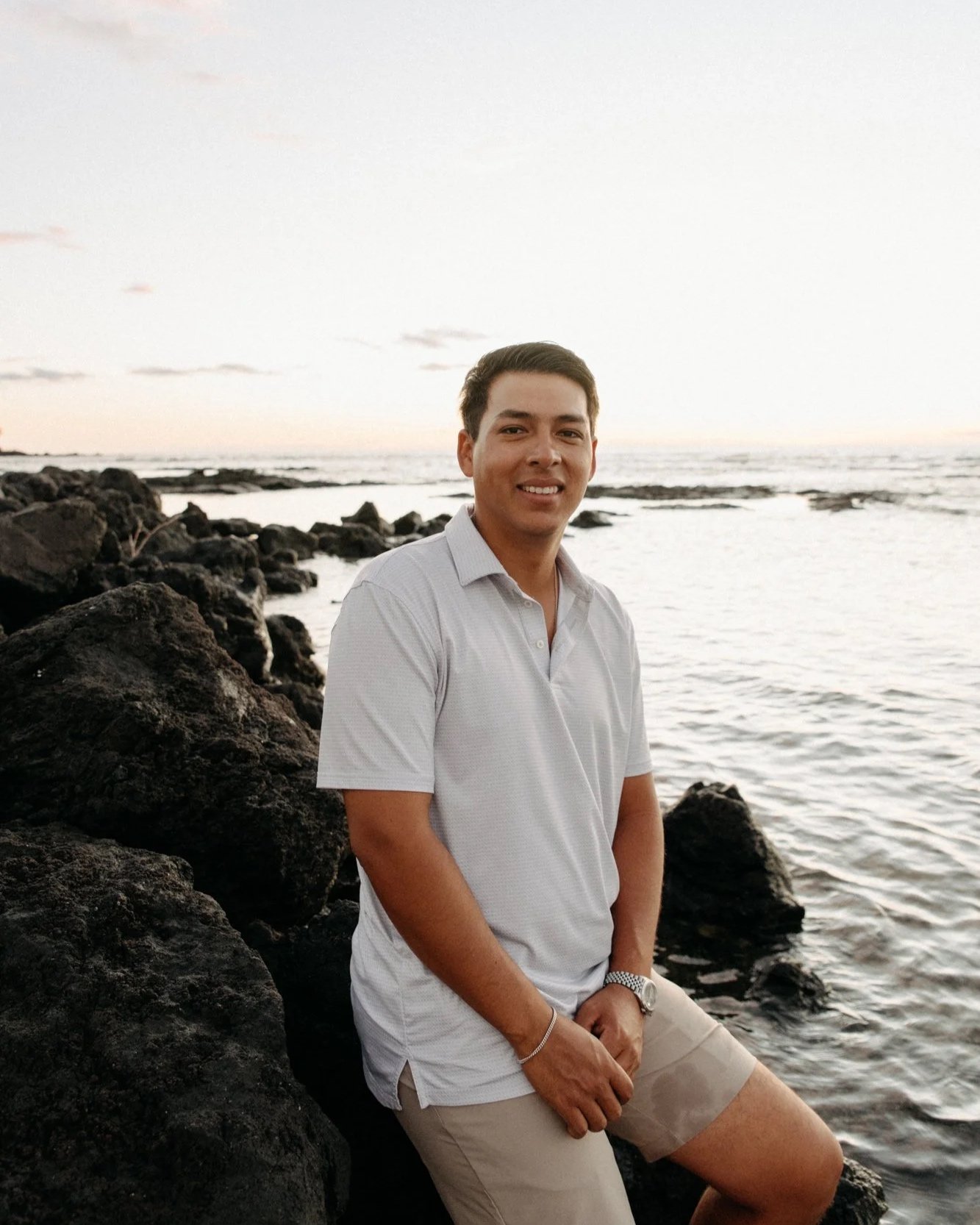 A young man in a white polo shirt and beige shorts sitting on rocks by the ocean during sunset, smiling at the camera.