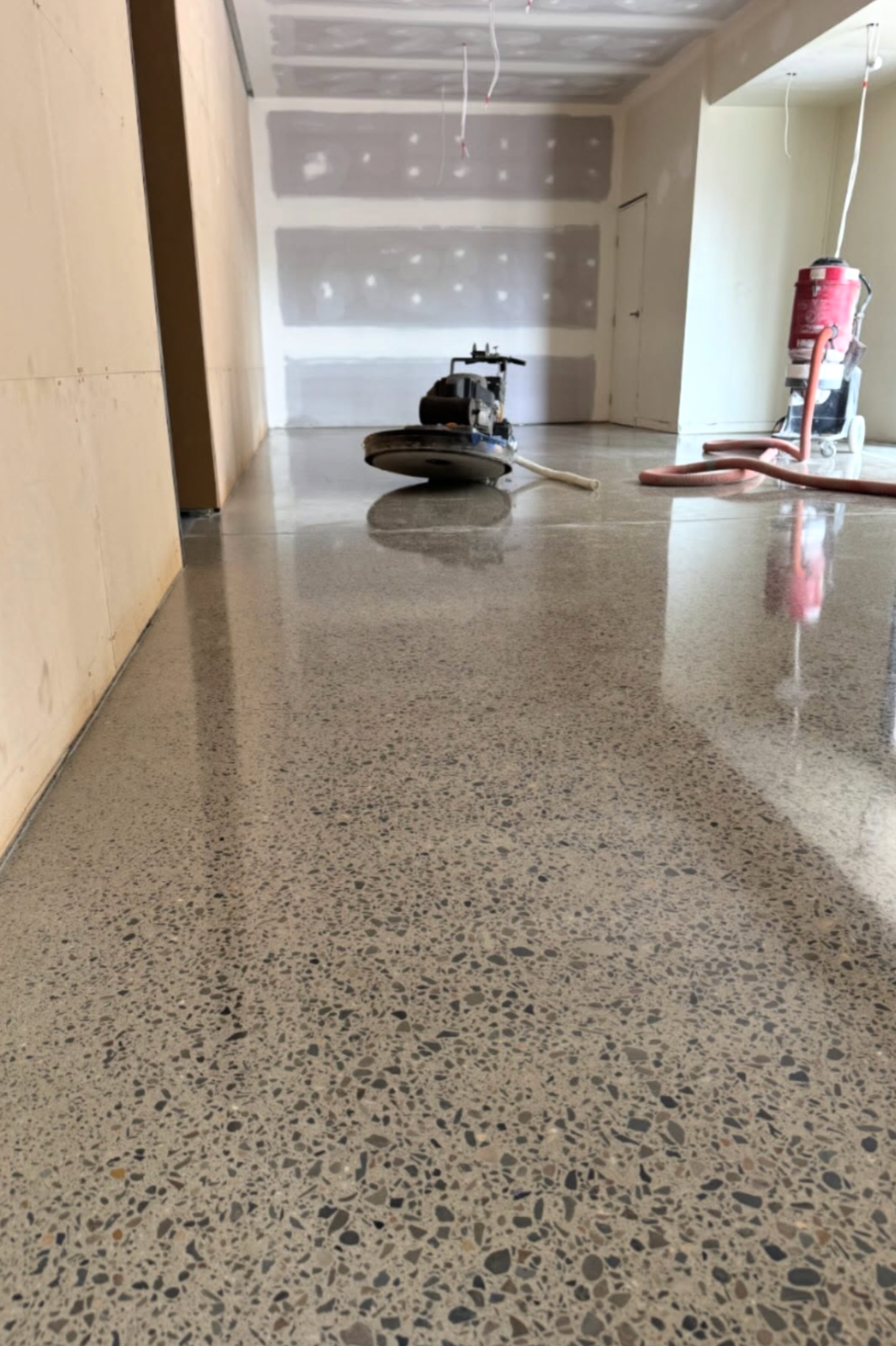 Construction site with polished terrazzo flooring, a power saw, and construction tools, walls with drywall in progress, and wires hanging from the ceiling.