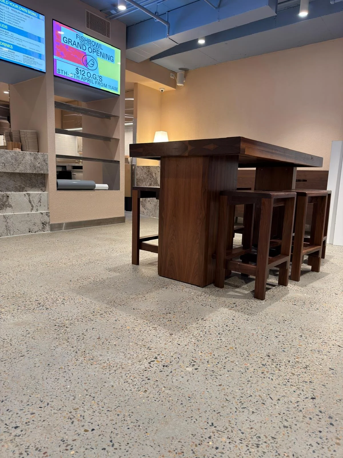 Indoor restaurant or cafe seating area with wooden tables and stools, digital menu signs on the wall, and ambient lighting.