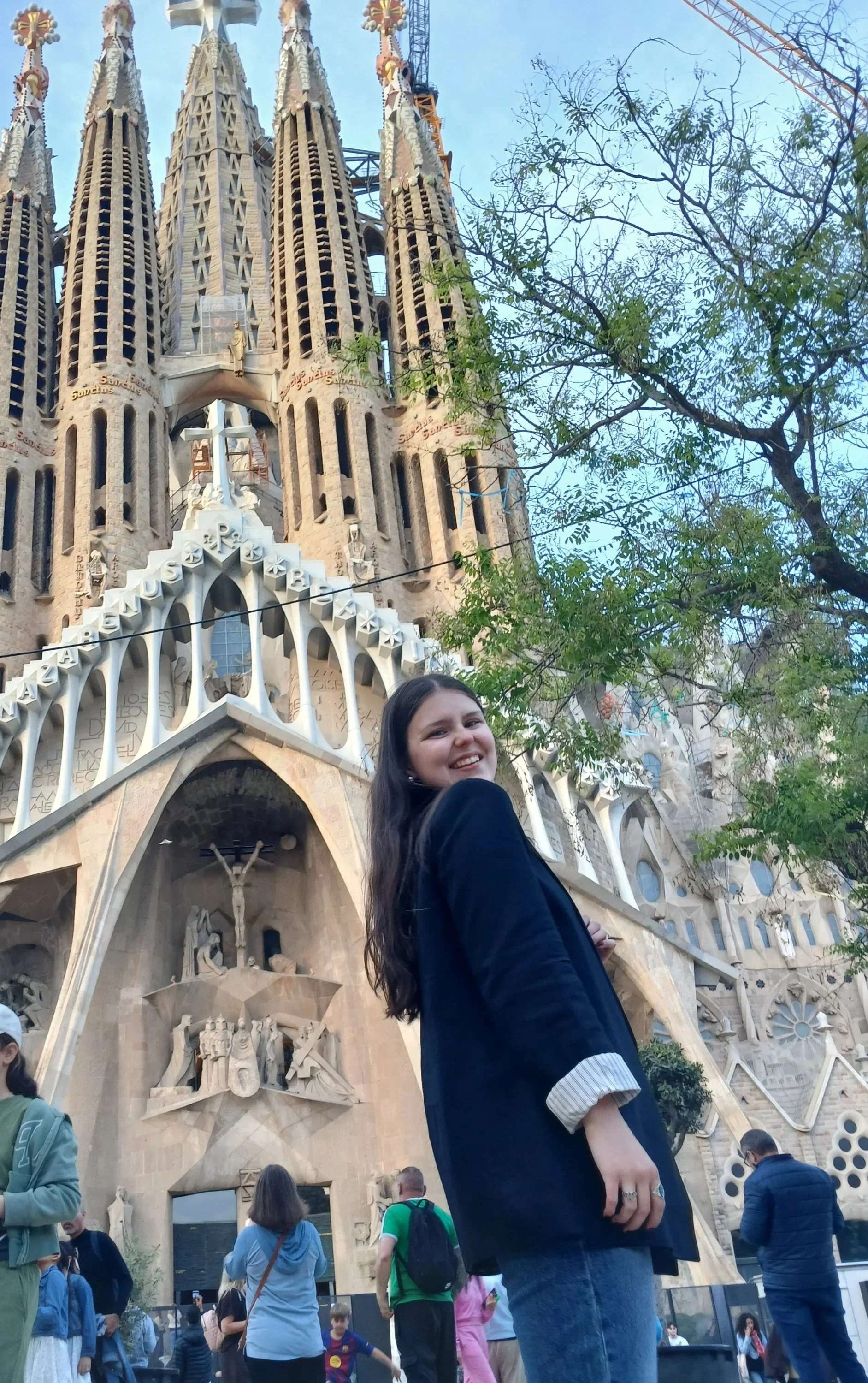 Joven sonriendo frente a la iglesia de la Sagrada Familia en Barcelona, con muchas personas alrededor y árboles en el fondo.