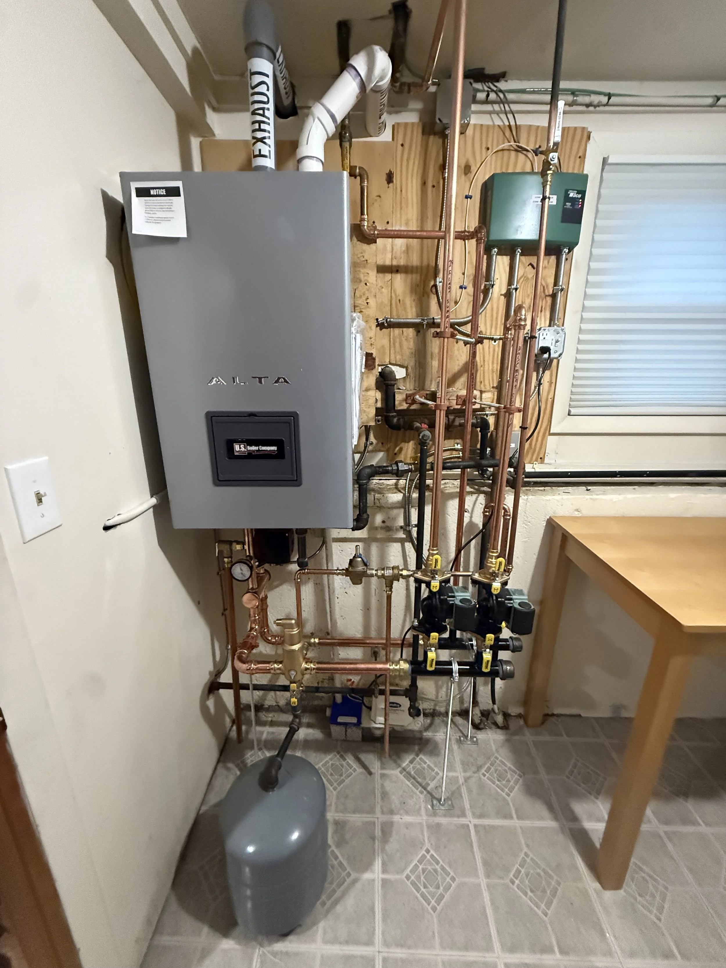 Heating and plumbing system installed in a small utility room with an oil tank, copper pipes, a tankless water heater, and a control box, near a window with a white shutter.