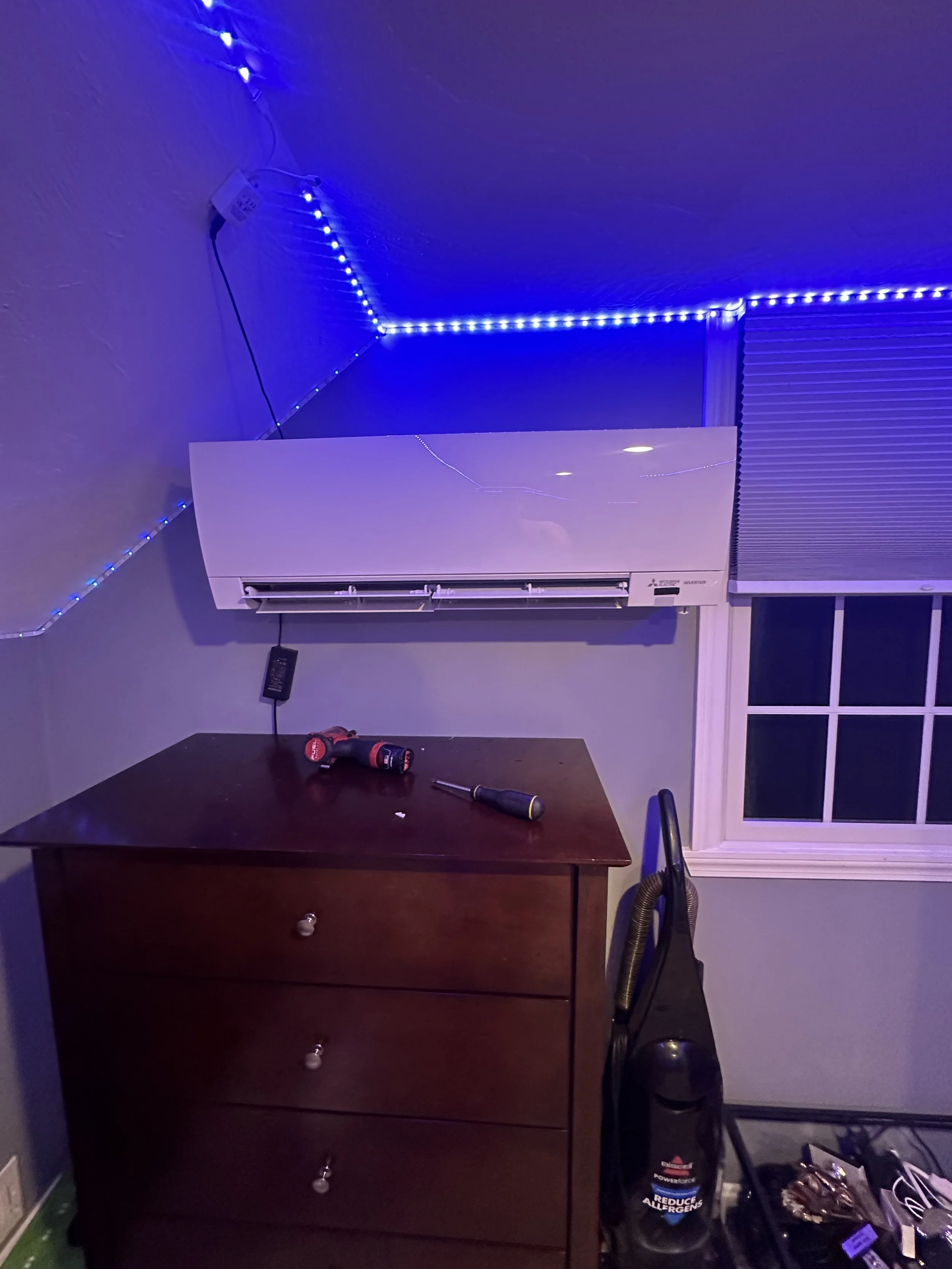 A room with purple LED strip lights along the ceiling, an air conditioning unit on the wall, a wooden dresser with three drawers, a cordless drill, a screwdriver, and a vacuum cleaner beside the dresser, a window with closed blinds, and a yellowish-green floor.