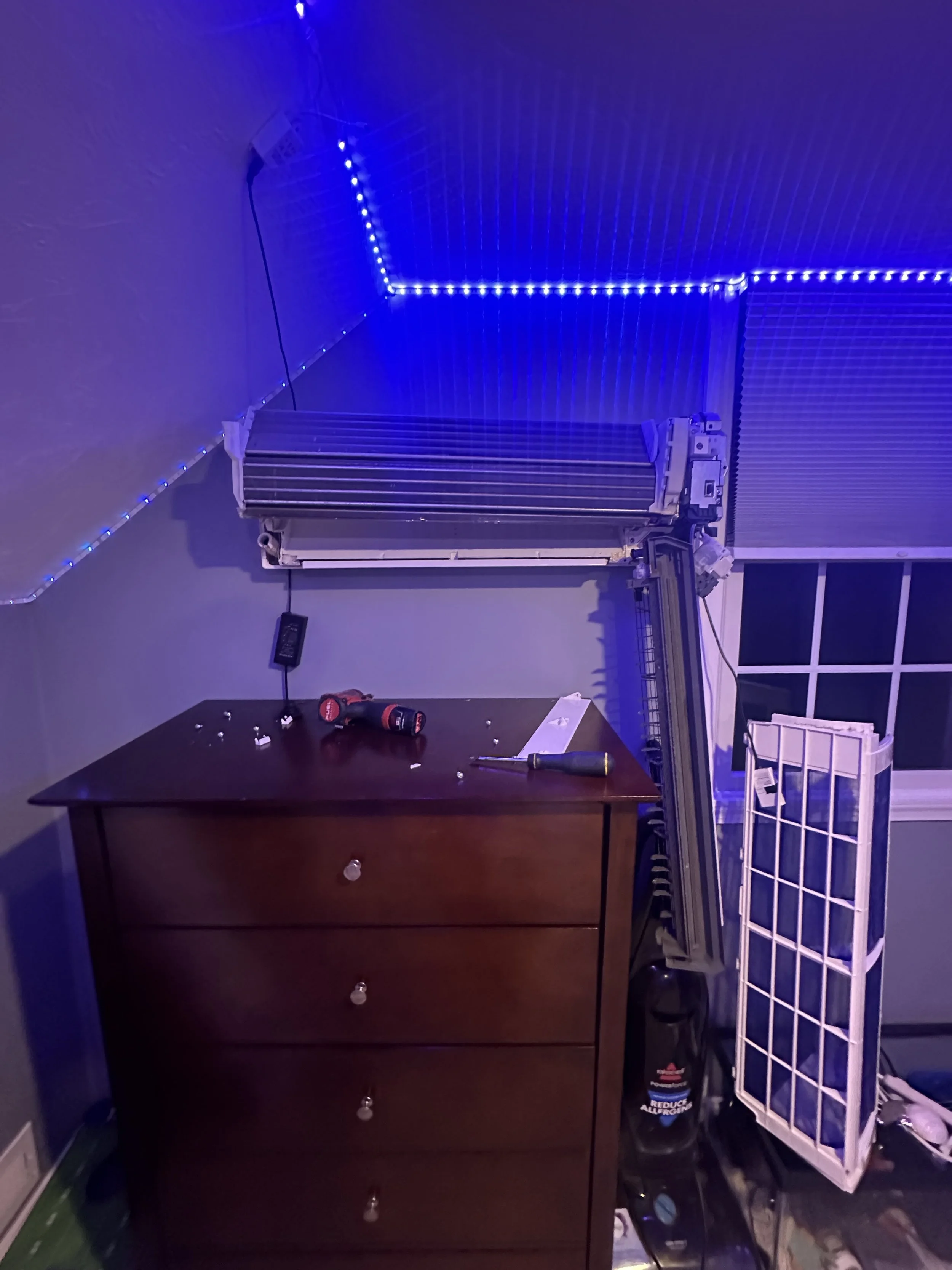 Room with 3-drawer dark brown wooden dresser, blue LED strip lights on the ceiling and wall, air conditioner unit, black power drill, small screws on the dresser, white wire shelving unit, window with black grid, vacuum cleaner, and various small items.