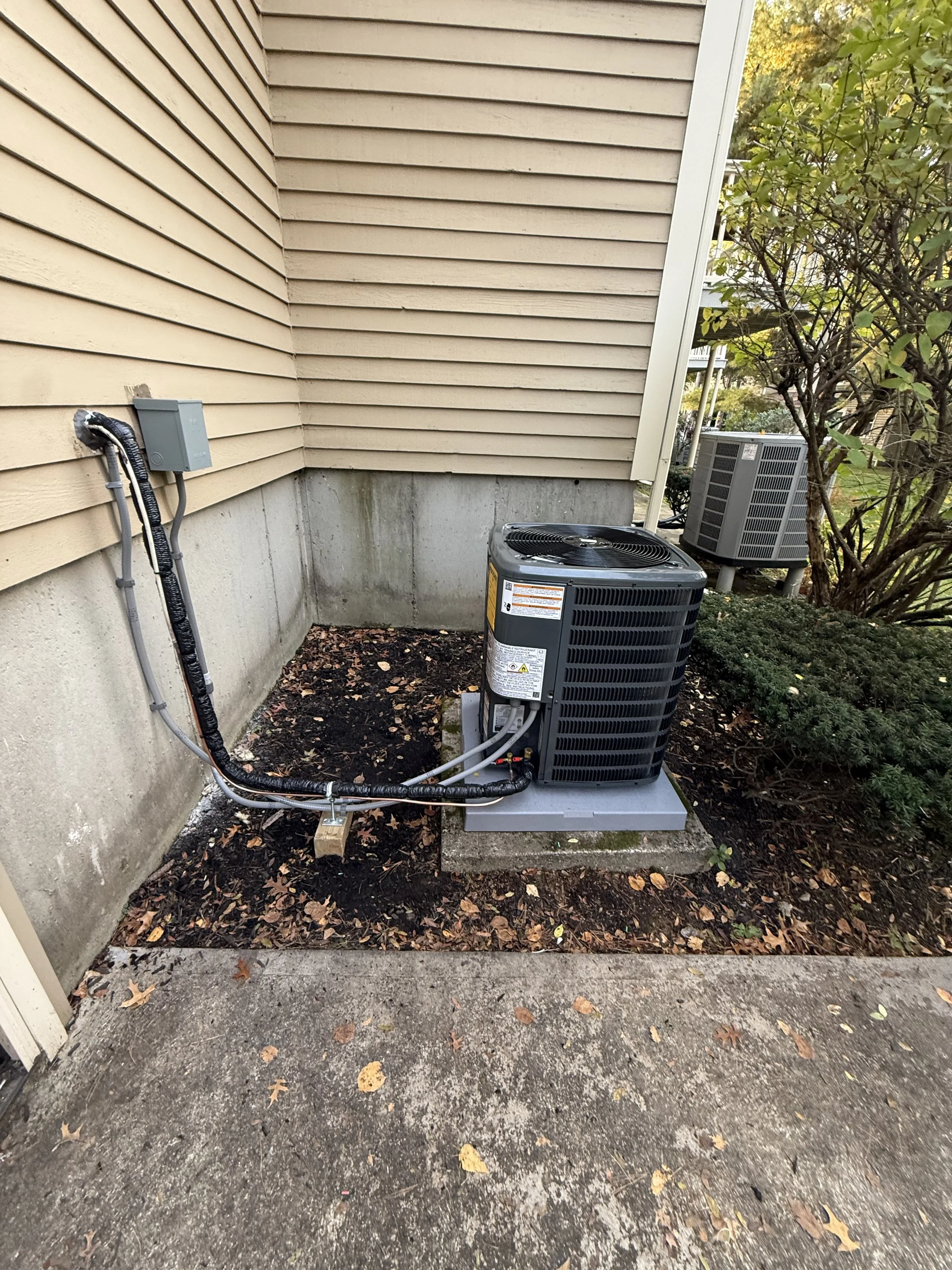 Outside view of an air conditioning unit installed beside a beige house with horizontal siding. The unit is on a concrete slab, surrounded by fallen leaves and dirt, with a bush and another air conditioning unit in the background.