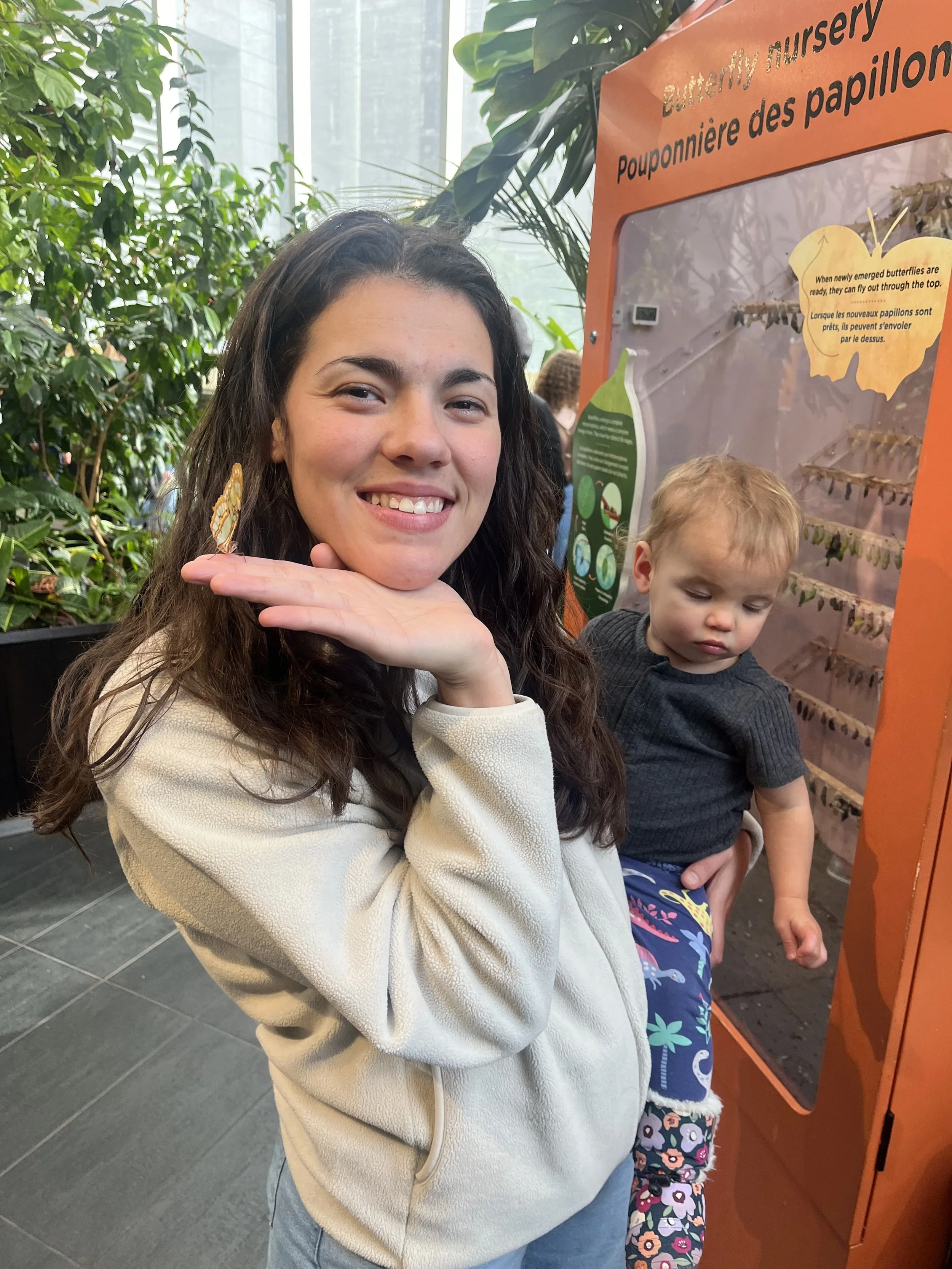 A smiling woman with long brown hair, wearing a cream-colored hoodie, holding her hand under her chin. Next to her is a young boy with blond hair, wearing a dark shirt and colorful shorts, looking down. They are in an indoor setting with green plants and an informational display about butterflies in the background.