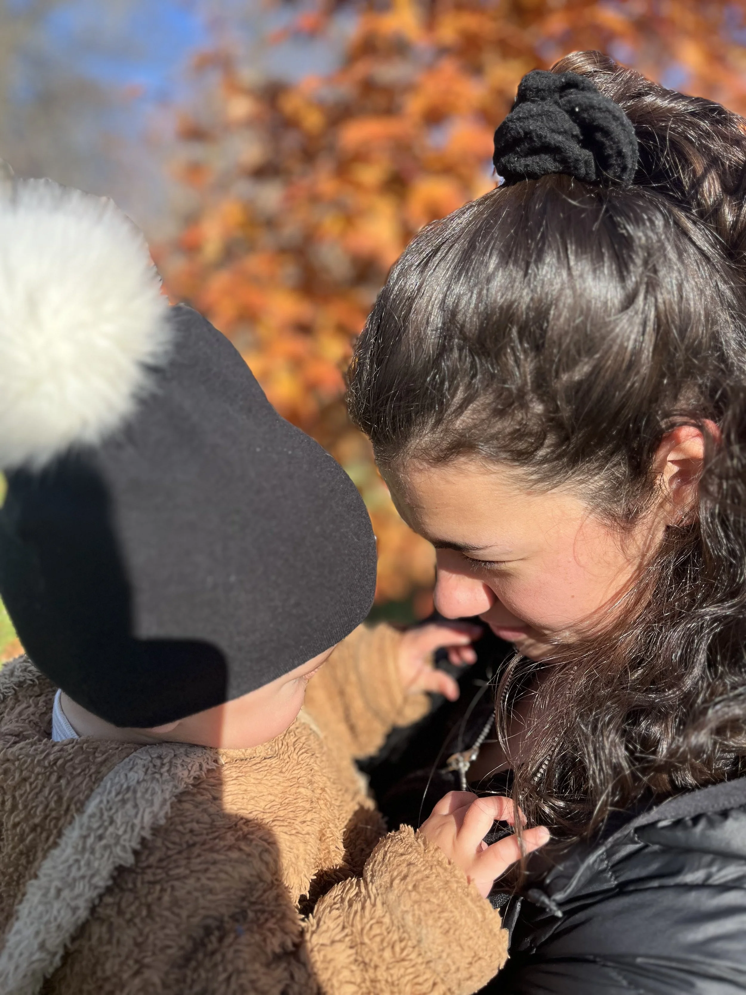A little boy and a woman facing each other closely outdoors with autumn leaves in the background, sharing an intimate moment.