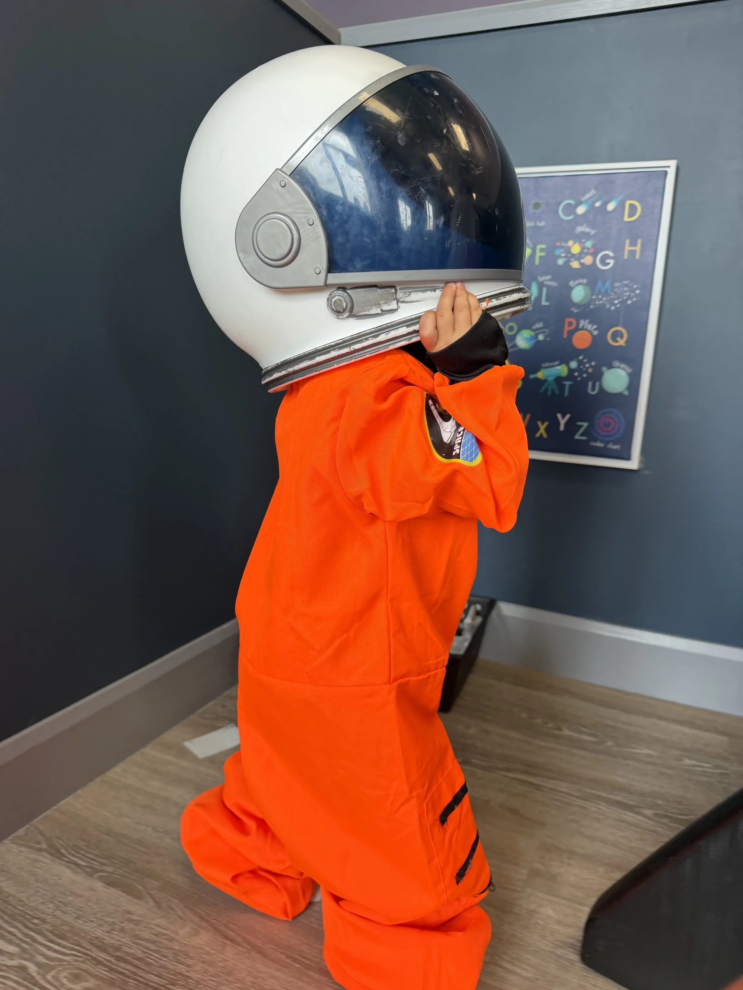 A child dressed in an orange astronaut costume with a space helmet, standing indoors in front of a dark wall and a poster with the alphabet and space-themed illustrations.