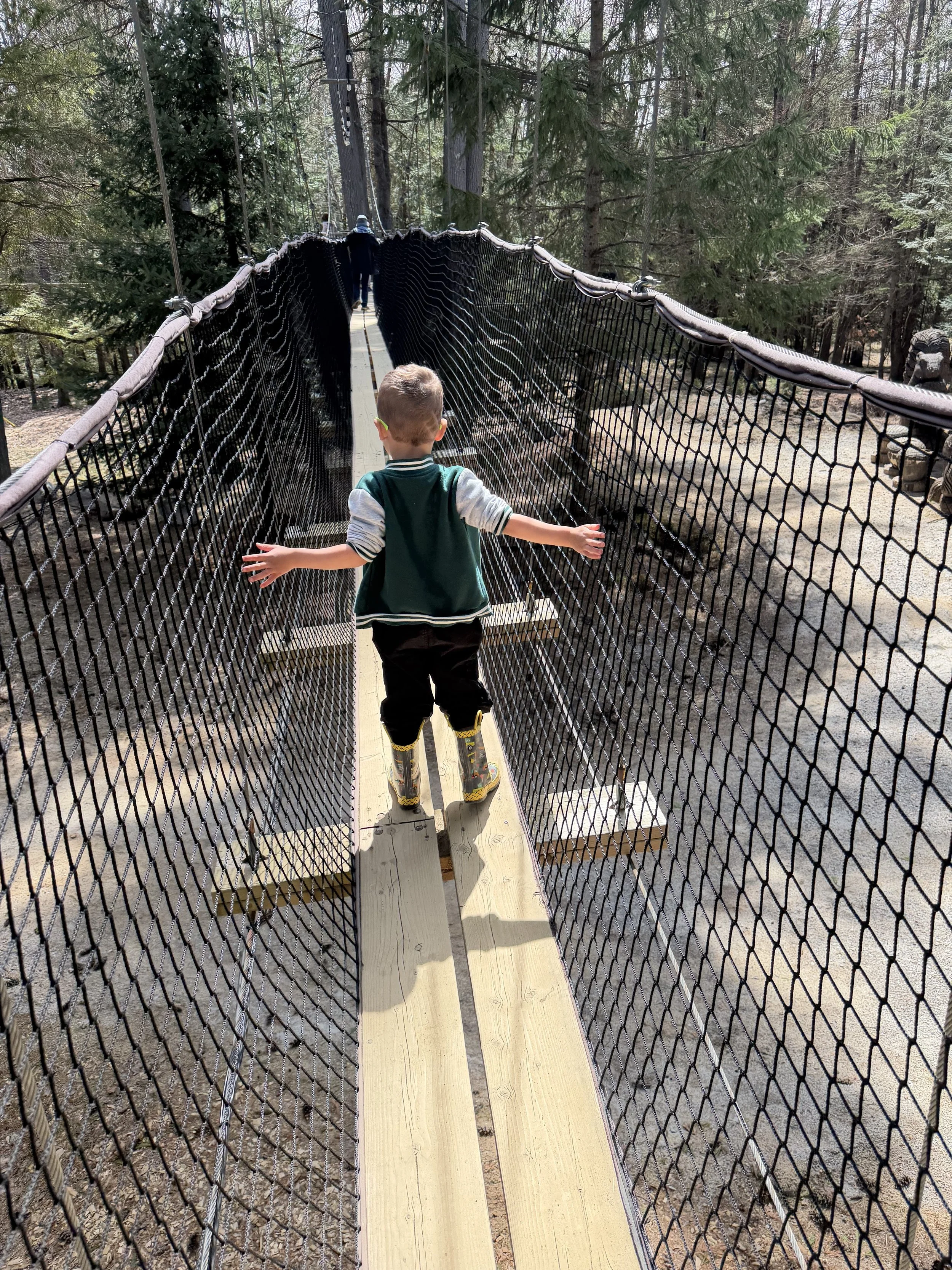 Child walking on a narrow wooden suspension bridge with black mesh sides in a forested area.