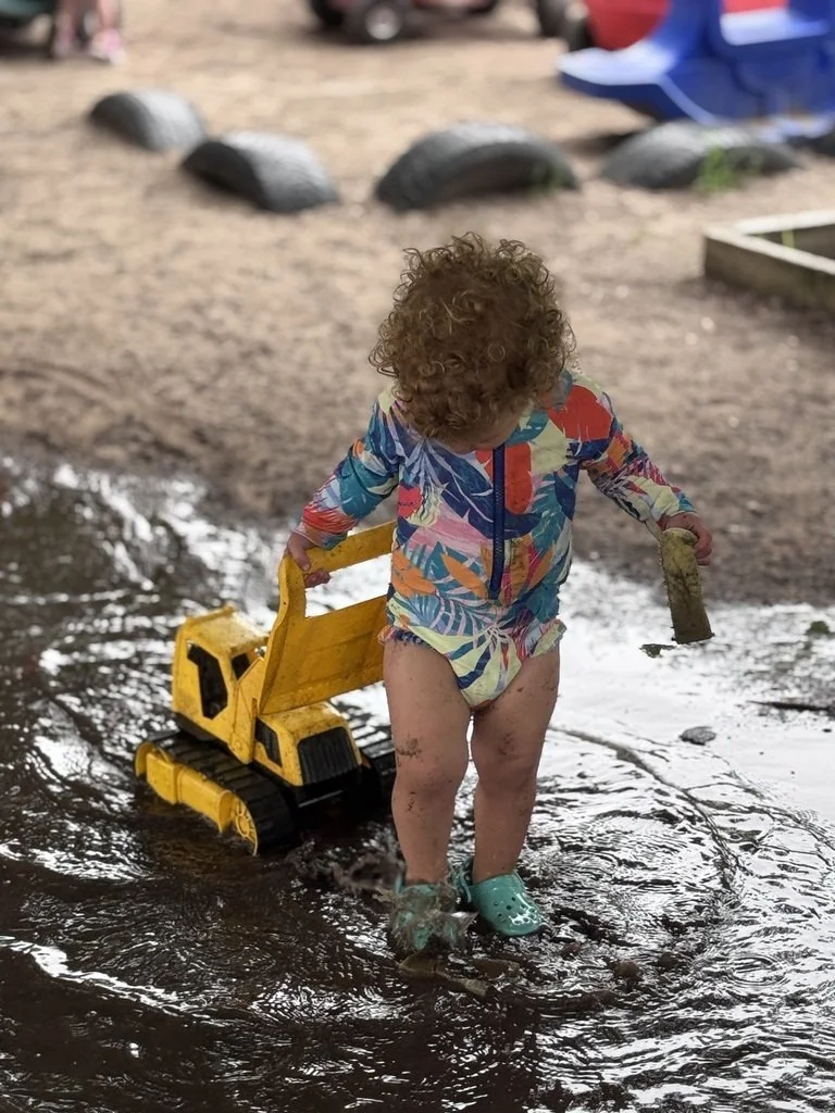 A young child with curly hair wearing a colorful patterned jacket and Crocs playing in a muddy puddle with a yellow toy excavator.