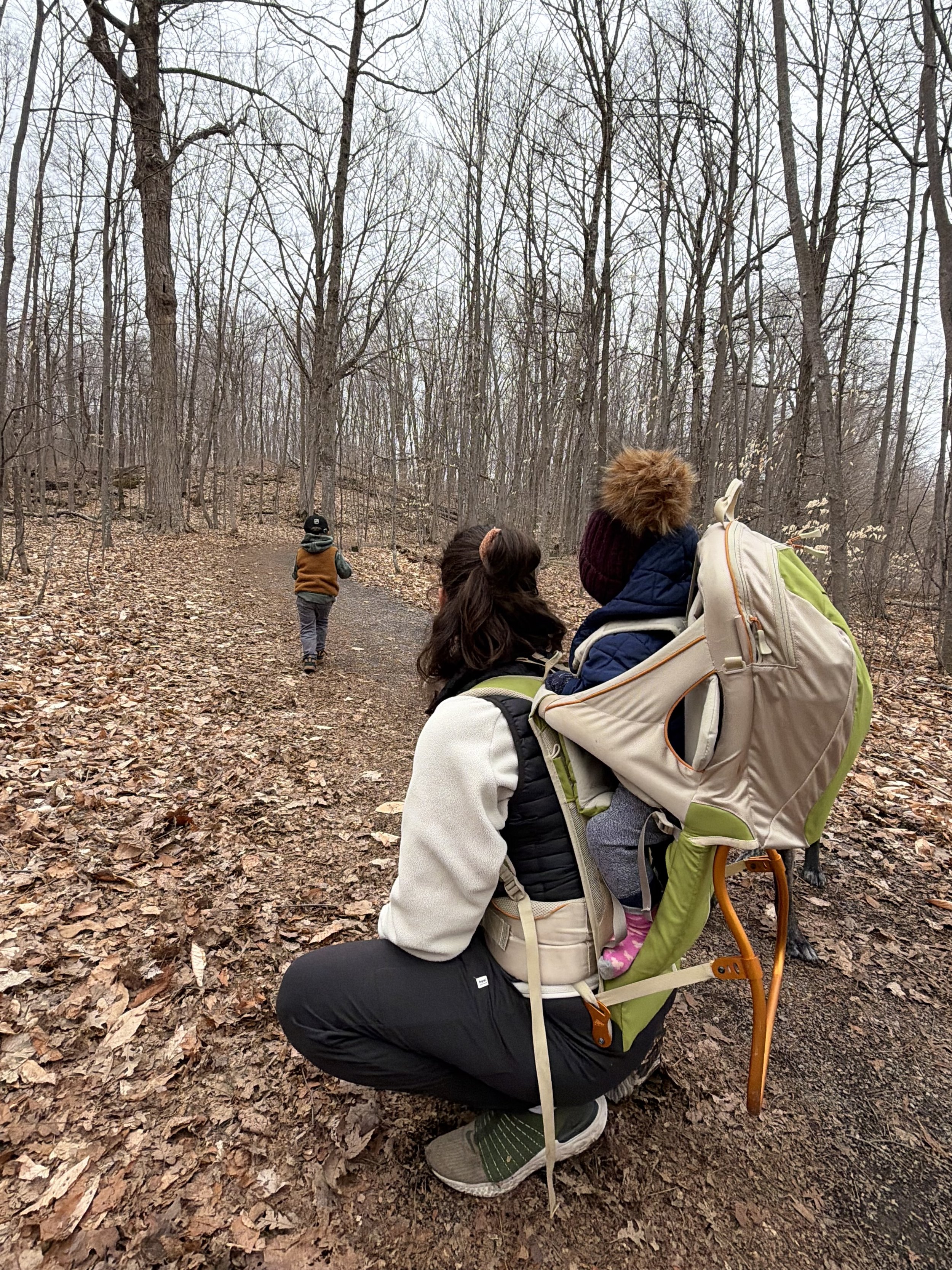 A woman crouching on a trail in a wooded area during fall, with fallen leaves on the ground and a backpack on her back. Other people are walking ahead on the trail, with leafless trees surrounding them.