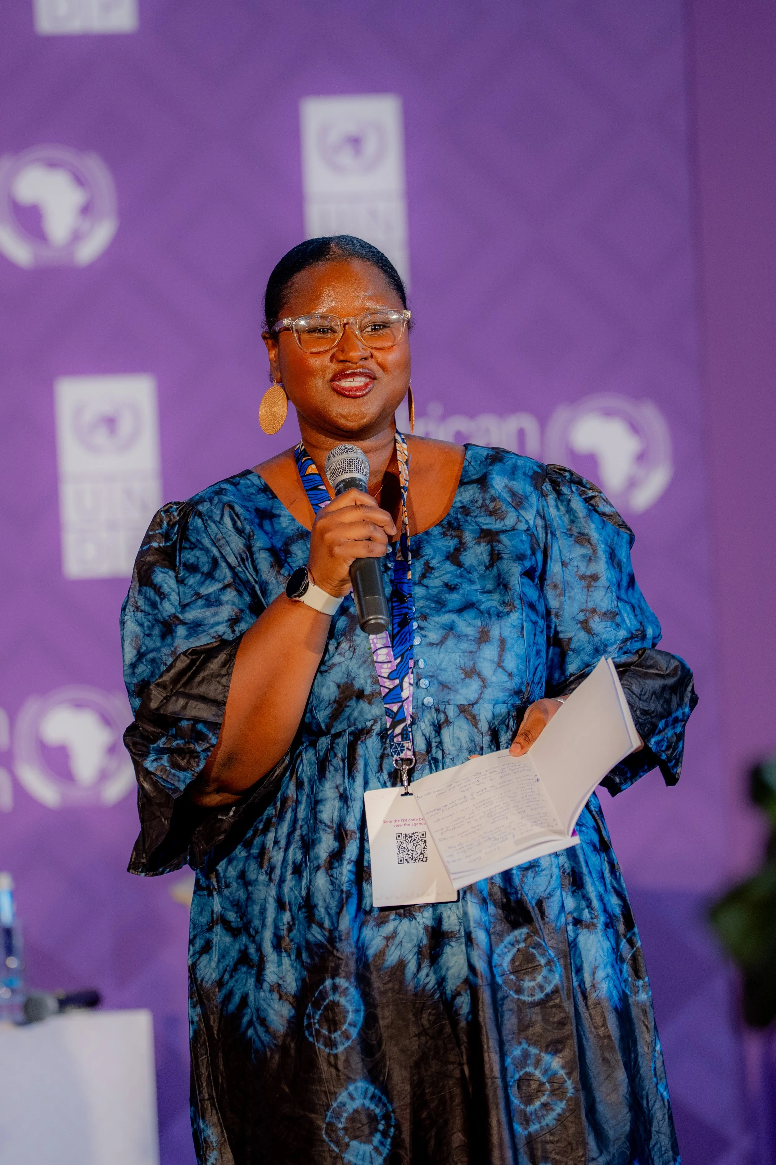 A woman speaking at a conference or event, holding a microphone and notes, with a purple backdrop featuring logos and flags of Africa in the background.
