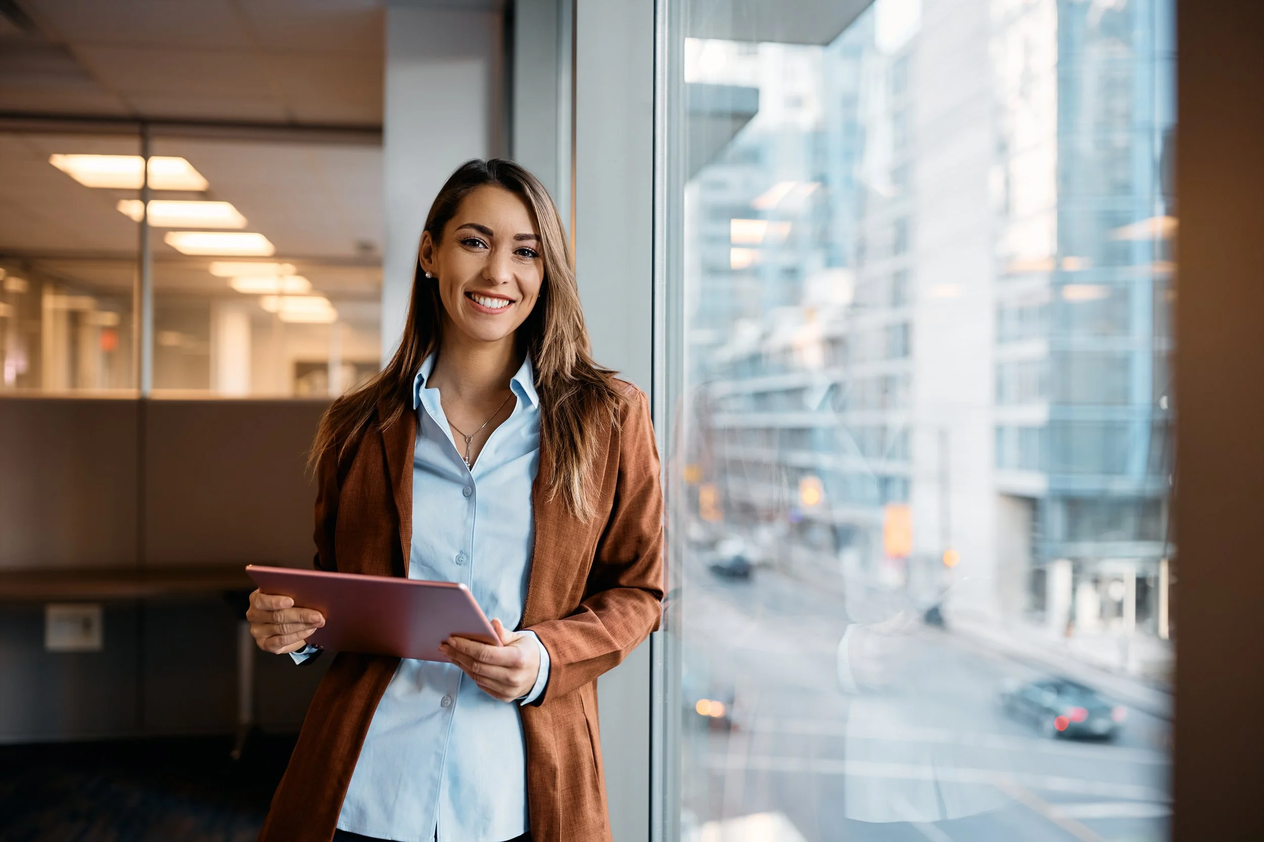 A woman dressed in business attire stands by a window in a modern office, holding a tablet and smiling at the camera, with city buildings visible outside.