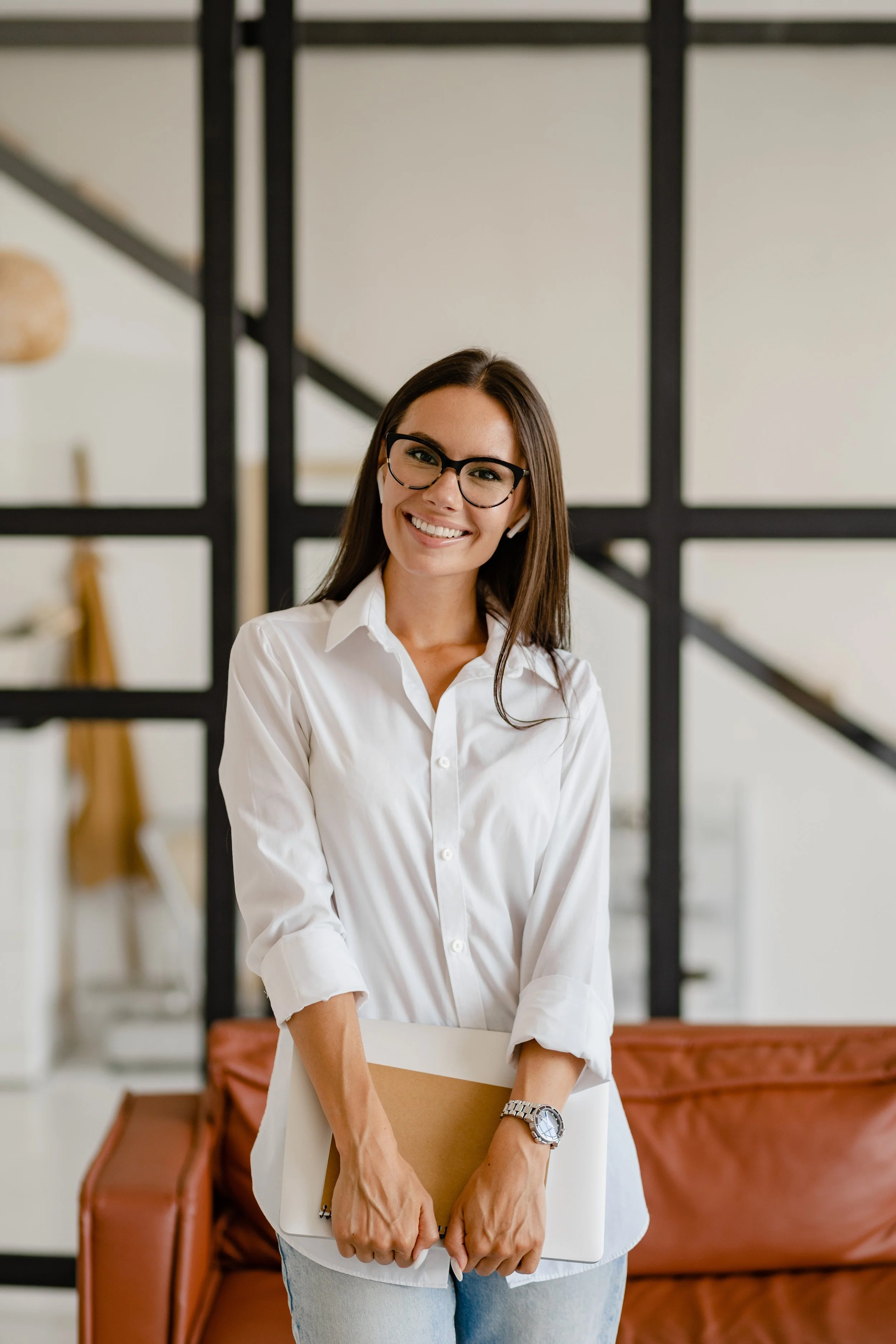 A woman with long dark hair, glasses, and a white shirt smiling and holding a folder and a laptop, standing in front of a sofa and a black geometric wall decor.