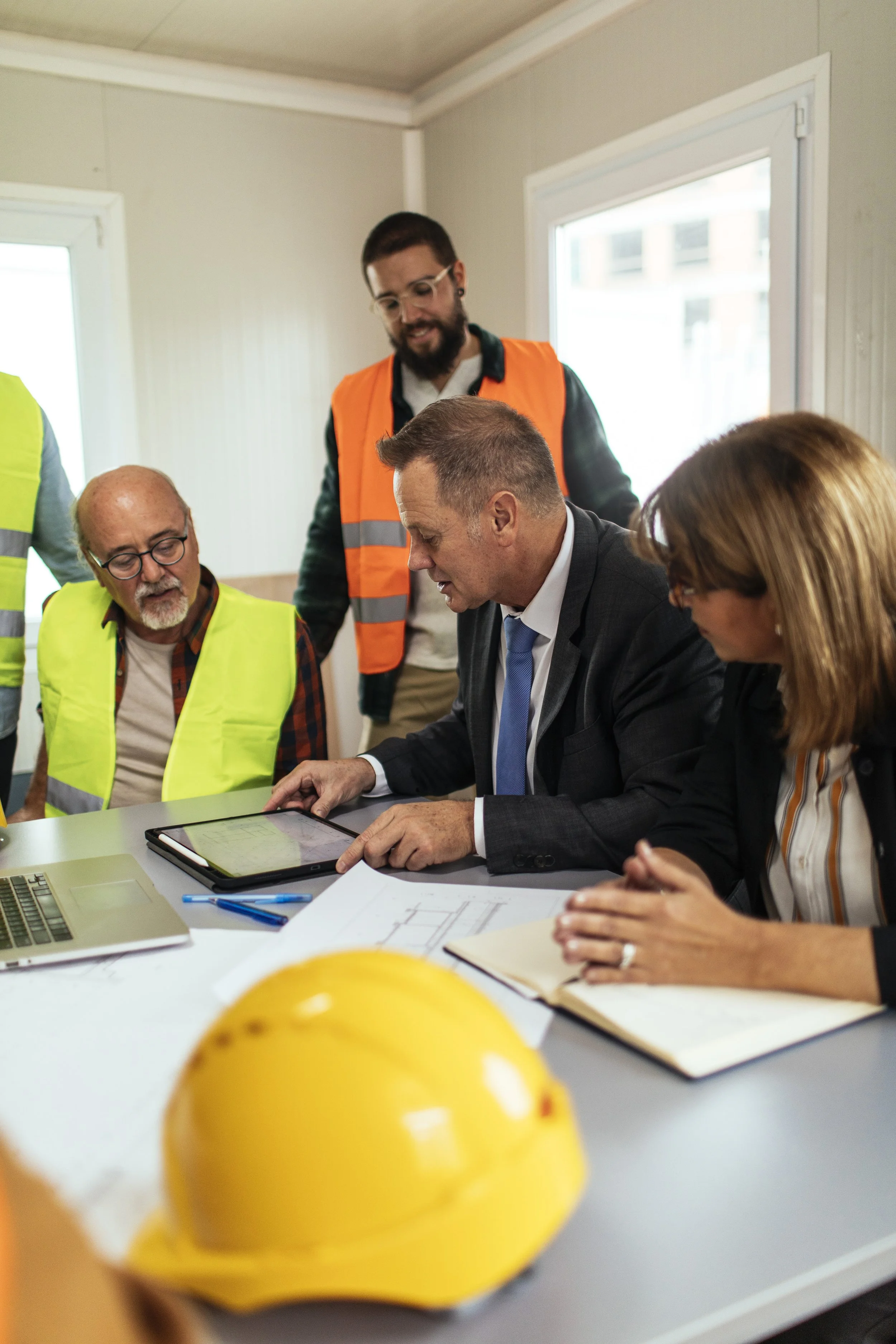 Group of professionals, some in safety vests and one in a suit, gathered around a table with documents, a laptop, and tablets, engaged in a discussion or meeting in a bright office. A yellow safety helmet is on the table in the foreground.