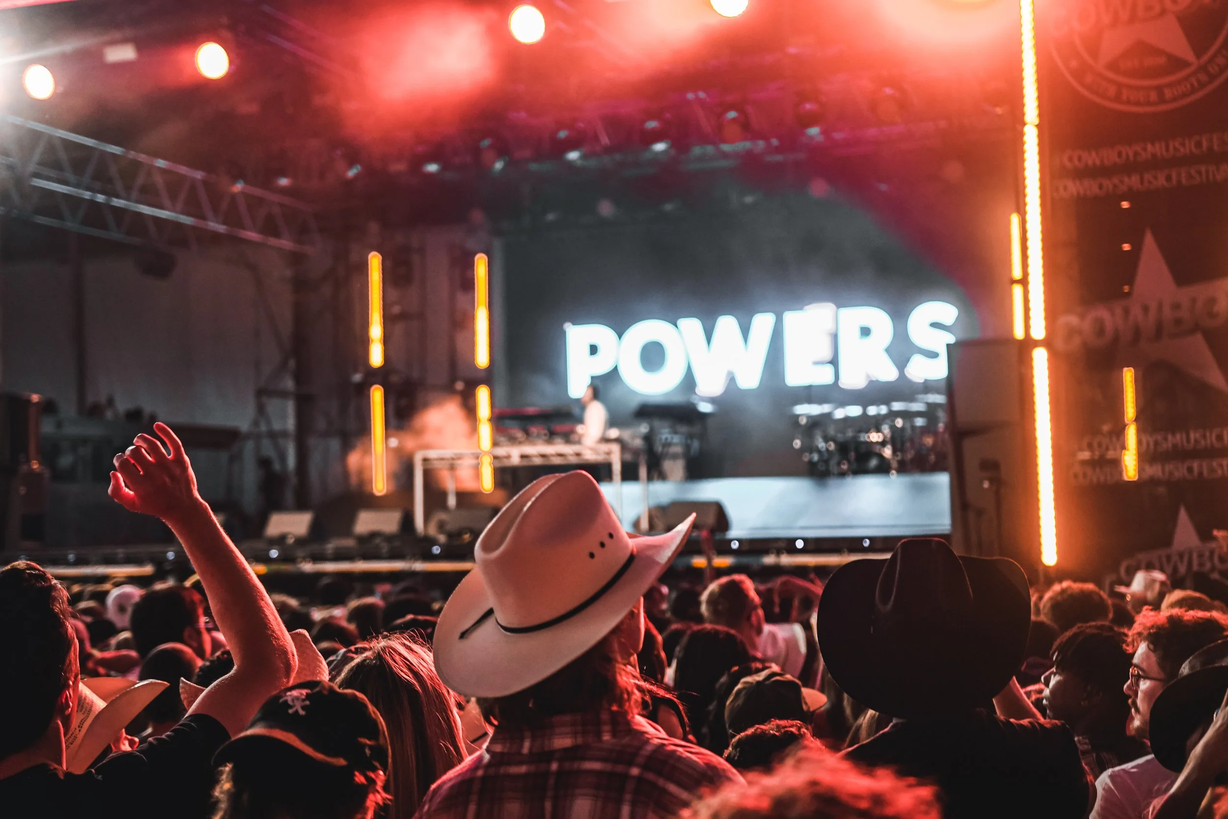 Concert crowd facing a stage with bright orange lights and a large illuminated sign that says 'POWERS.' The audience includes people wearing cowboy hats, with one arm raised in the foreground.