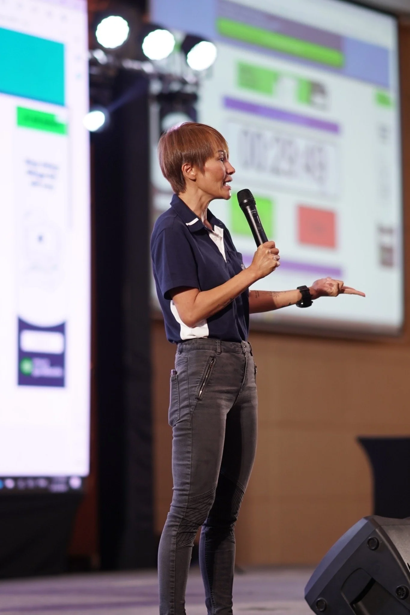 A woman with short reddish-brown hair standing on a stage, speaking into a microphone. She is wearing a dark polo shirt and gray pants with zipper details. In the background, there's a large screen with colorful graphics and text.