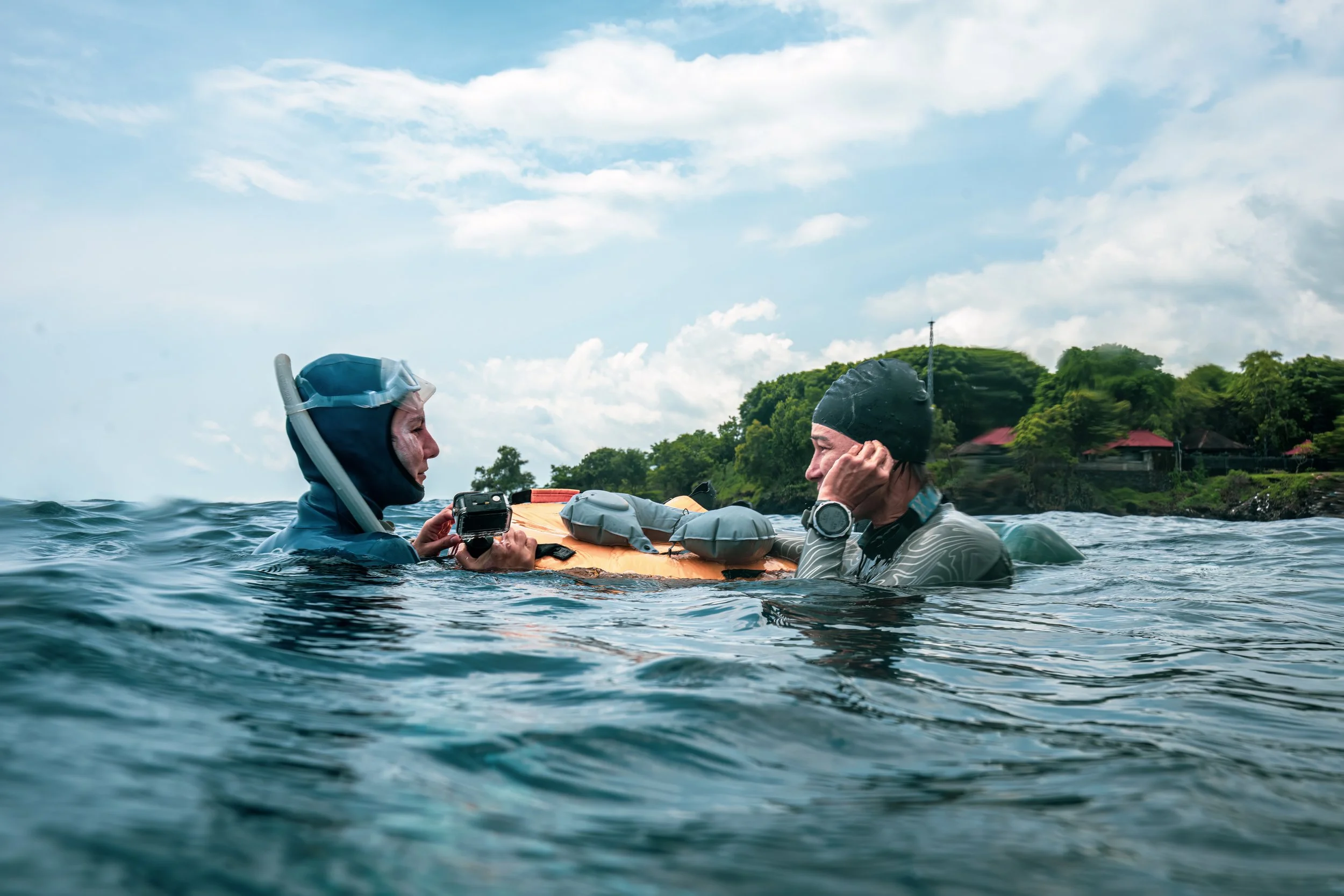 Two people in the water, one holding a camera, with a partially submerged surfboard or flotation device, and a shoreline with trees and houses in the background.