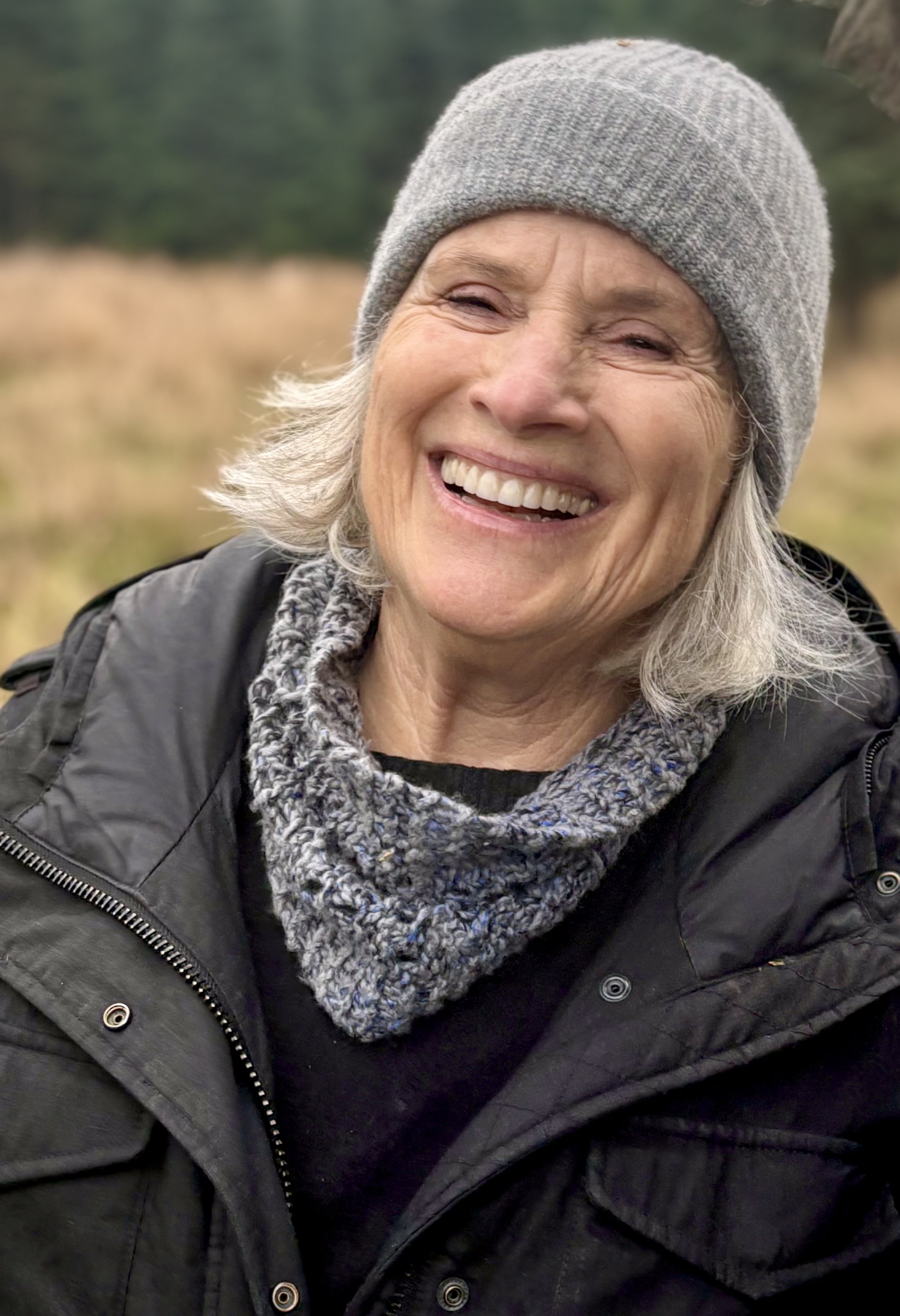 Close-up of a smiling older woman with gray hair, wearing a gray knit beanie, a black jacket, and a gray scarf outdoors.