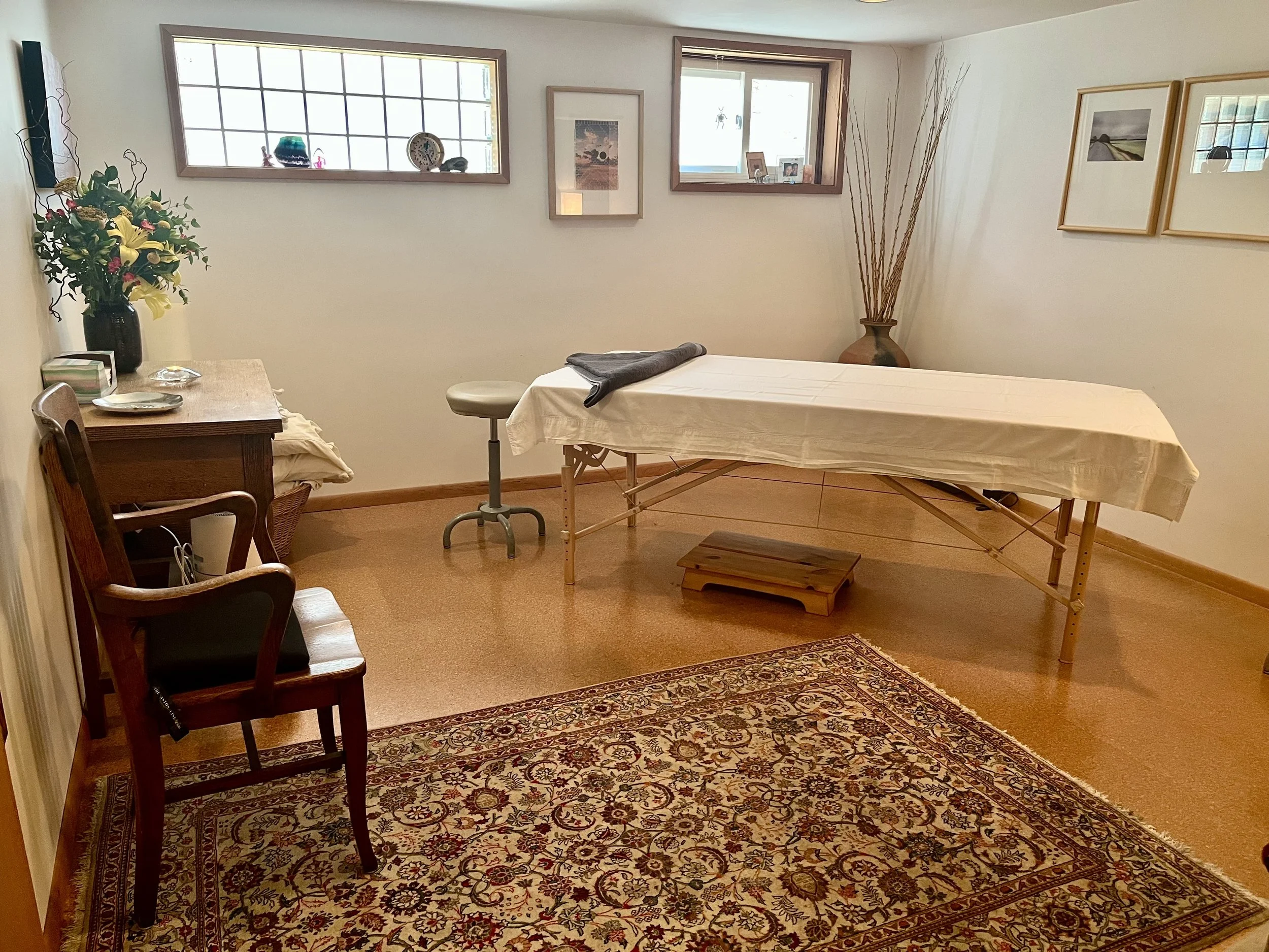 A room set up as a massage or therapy space with a massage table covered in a white sheet, a folded gray towel, and small stool. There is a large, patterned area rug, a wooden side table with a vase of flowers, and framed artwork on the walls. Two small windows with wood trim let in natural light, and a decorative large ceramic vase with tall dried branches is in the corner.
