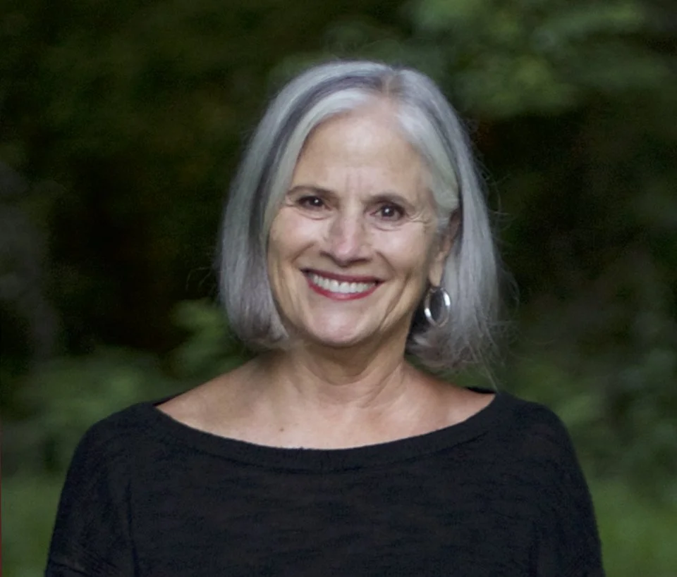 A smiling woman with shoulder-length gray hair, wearing a black top and hoop earrings, standing outdoors with blurred greenery in the background.