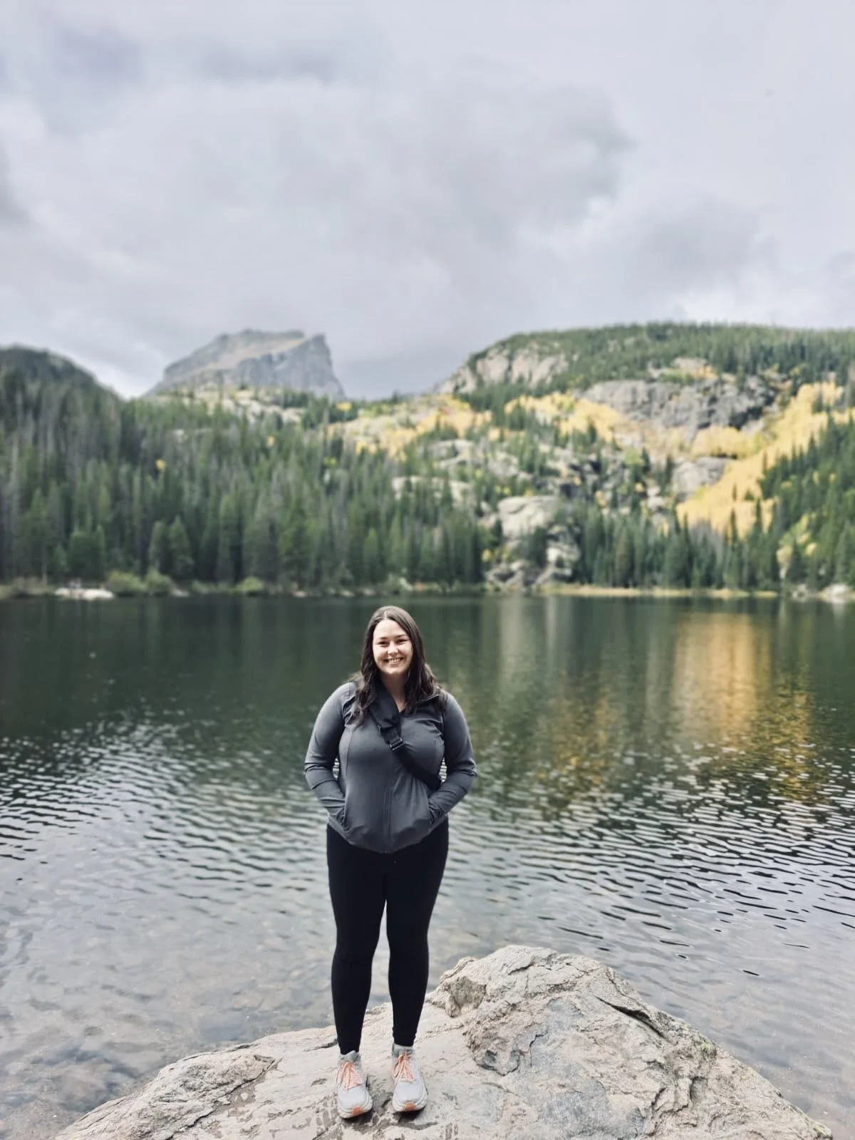 A woman standing on a rock by a lake with green forested mountains and cliffs in the background, under cloudy skies.