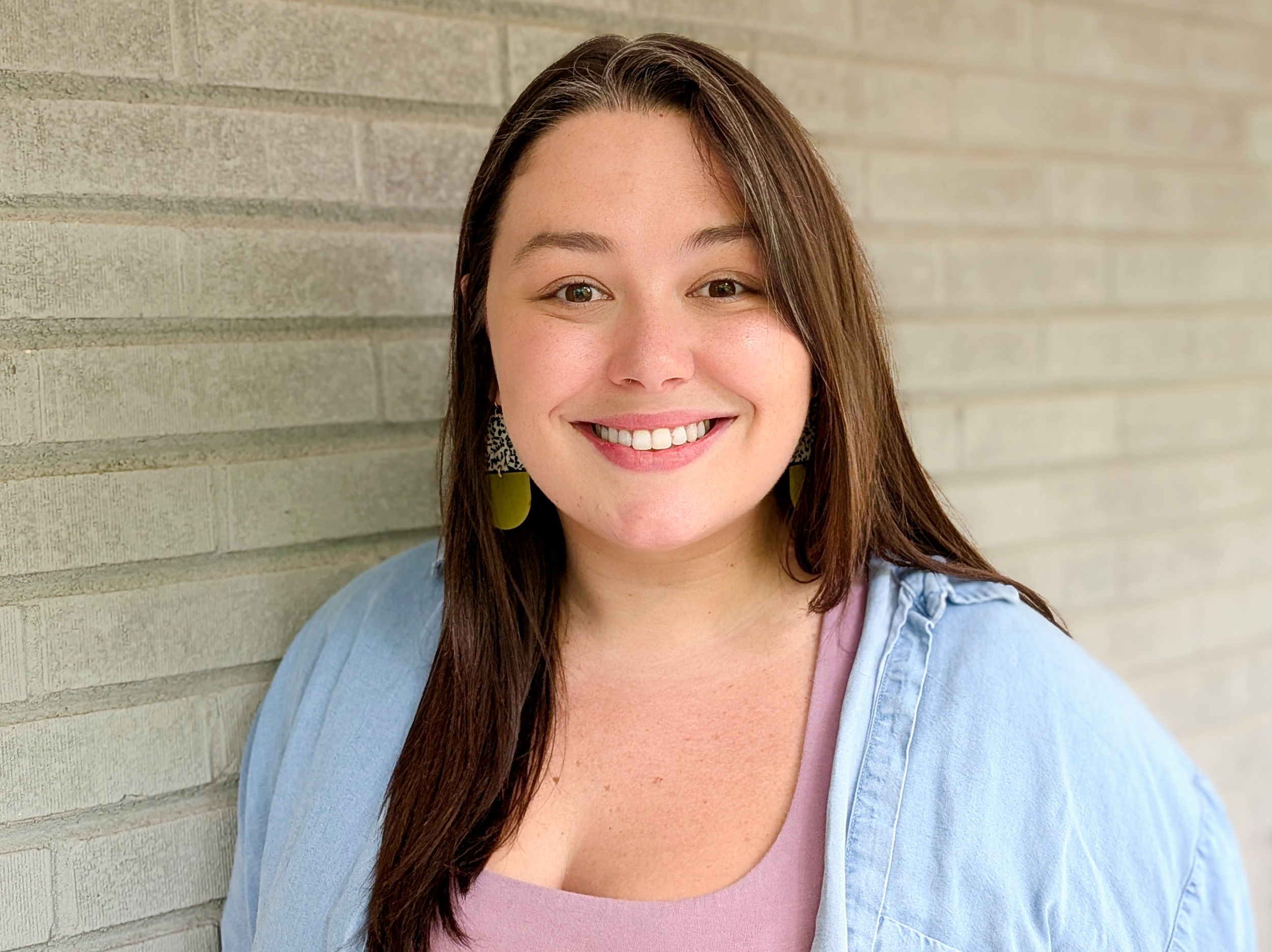 A close-up of a young woman smiling, with long brown hair, wearing earrings, standing against a beige brick wall.