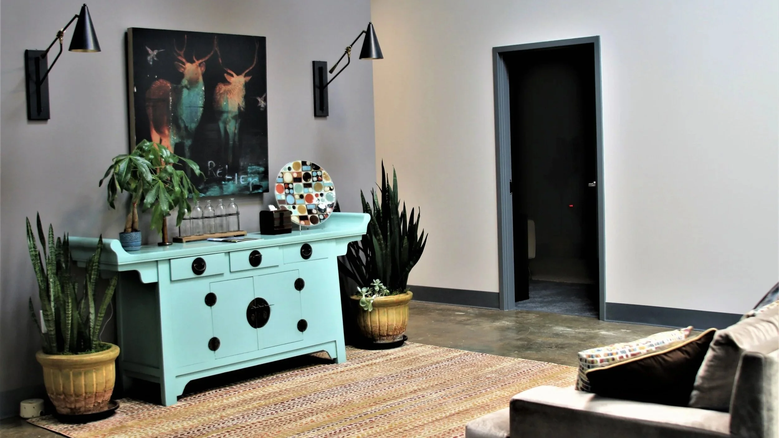 A modern interior room with a light blue sideboard, potted plants, wall sconces, and artwork, with an open doorway leading to another room.