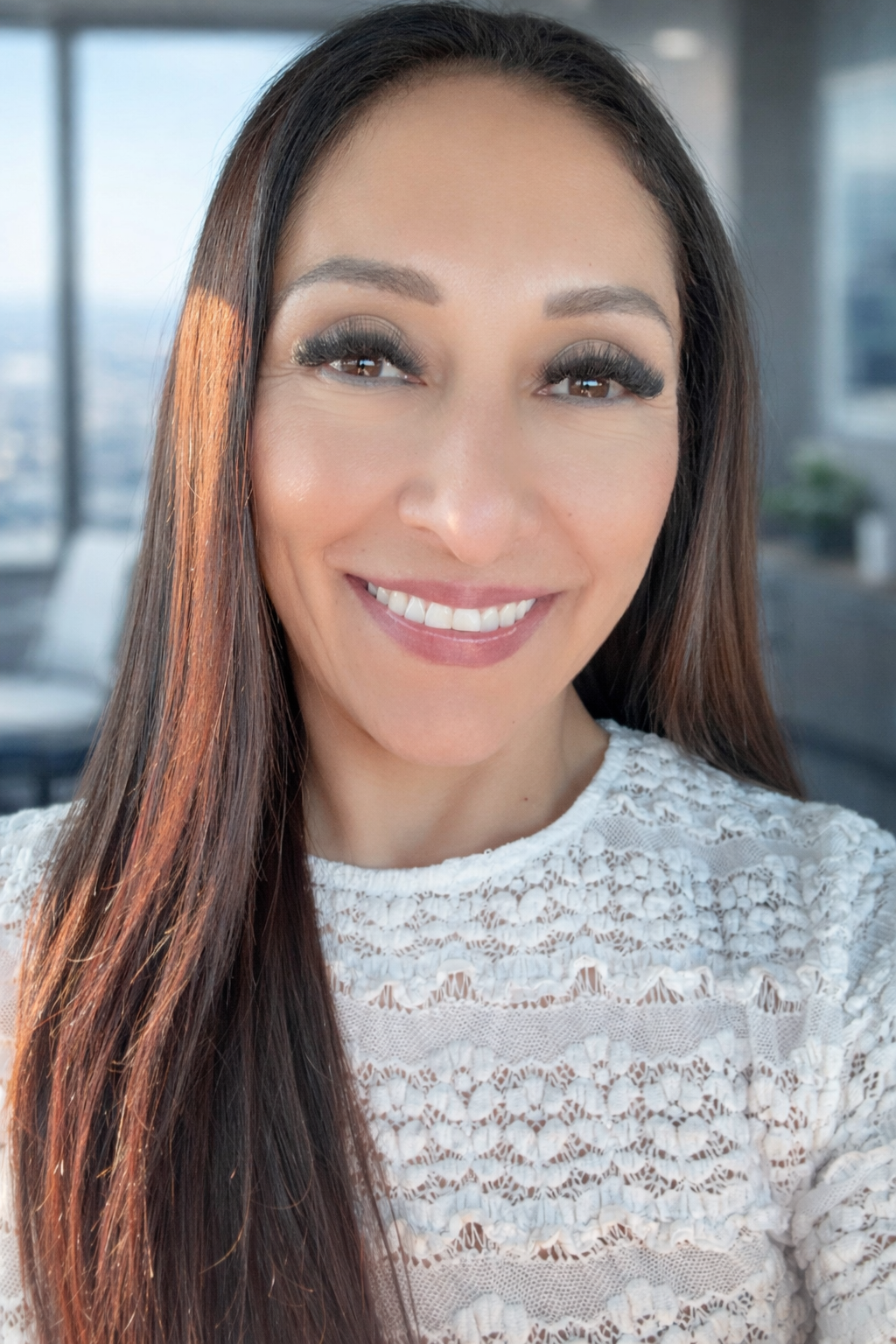 A woman with long dark hair smiling, wearing a white lace top, indoors with a city view in the background.