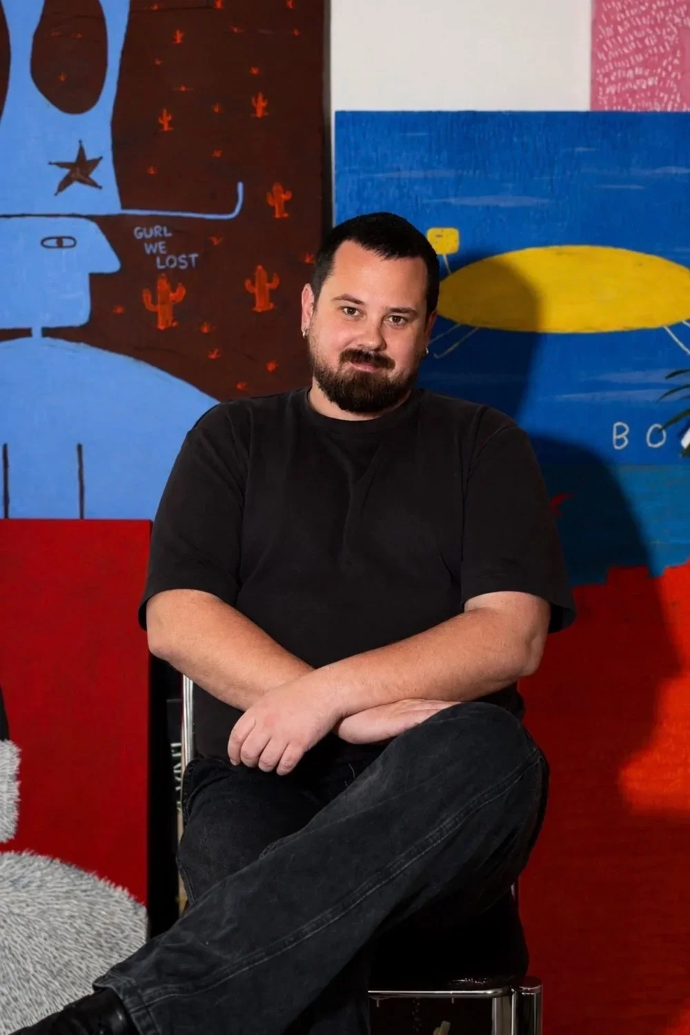 A man with dark hair and a beard, wearing a black shirt, sitting with one leg crossed over the other in front of colorful abstract wall art.