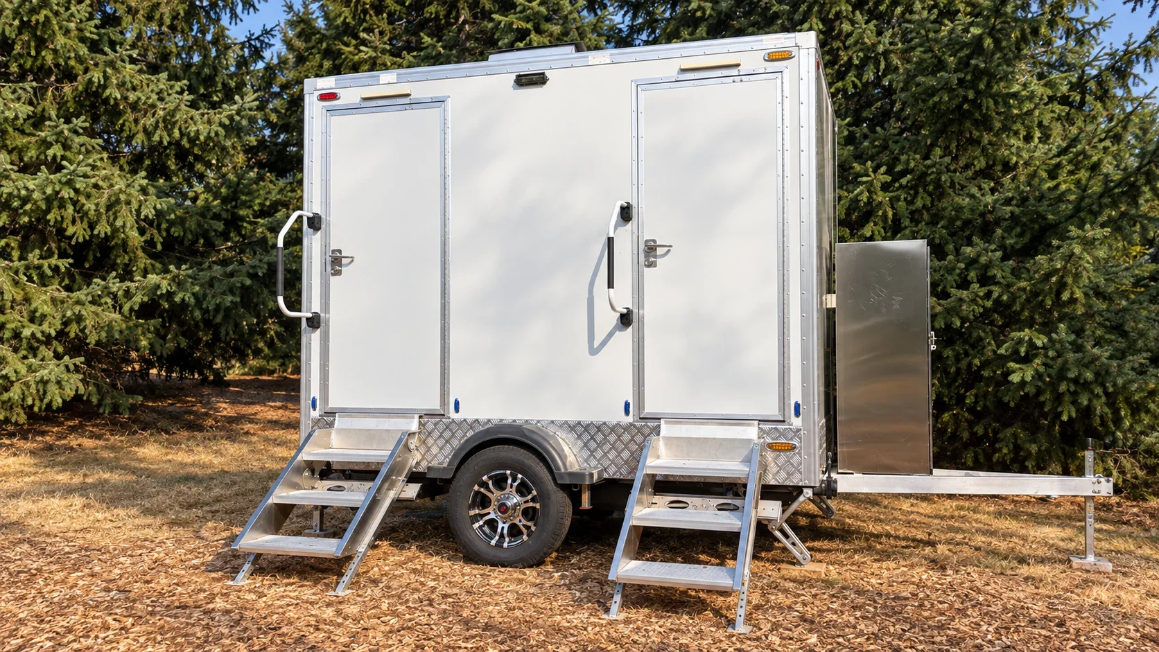 White two-room restroom trailer with separate entrances and aluminum stairs in a wooded outdoor setting.